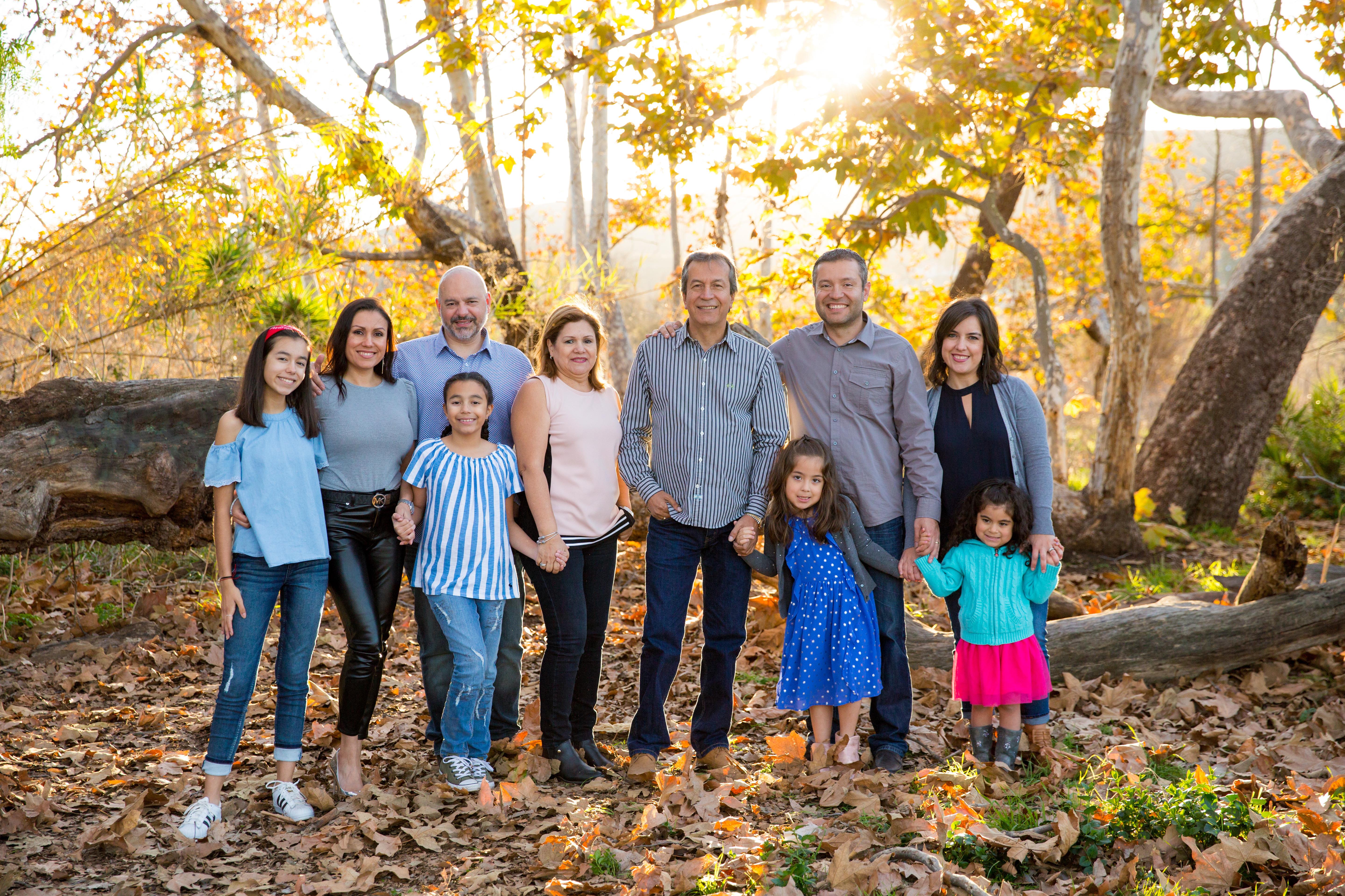 Family stands together in a park with trees and fallen leaves under bright sunlight.