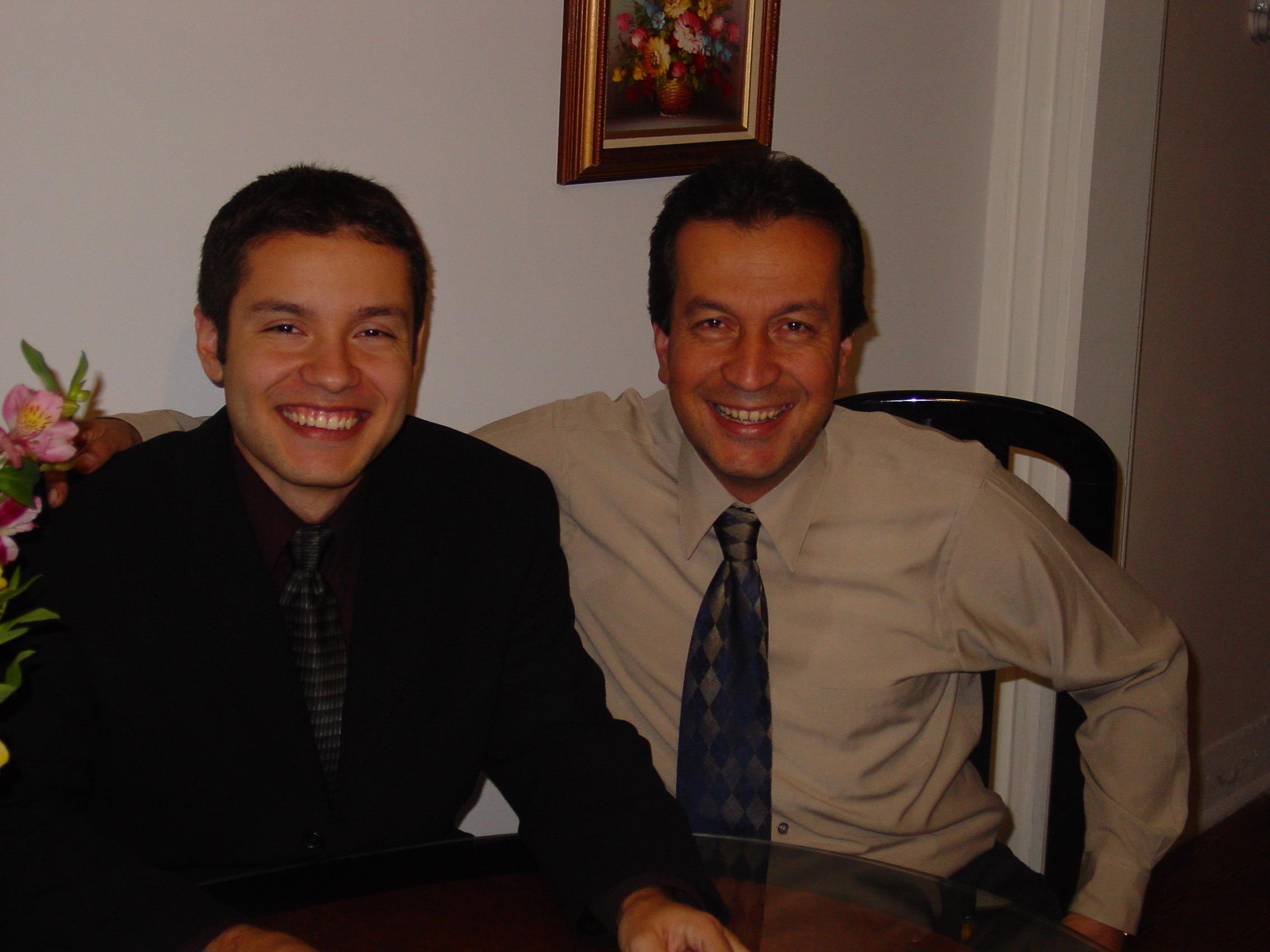 Two men share a moment at a table in a room with a light-colored wall and artwork.