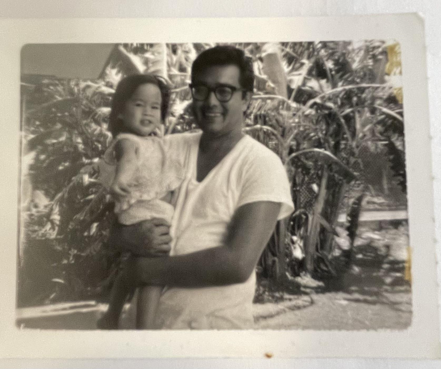 A father holds his young daughter in a garden filled with plants. They look happy together.