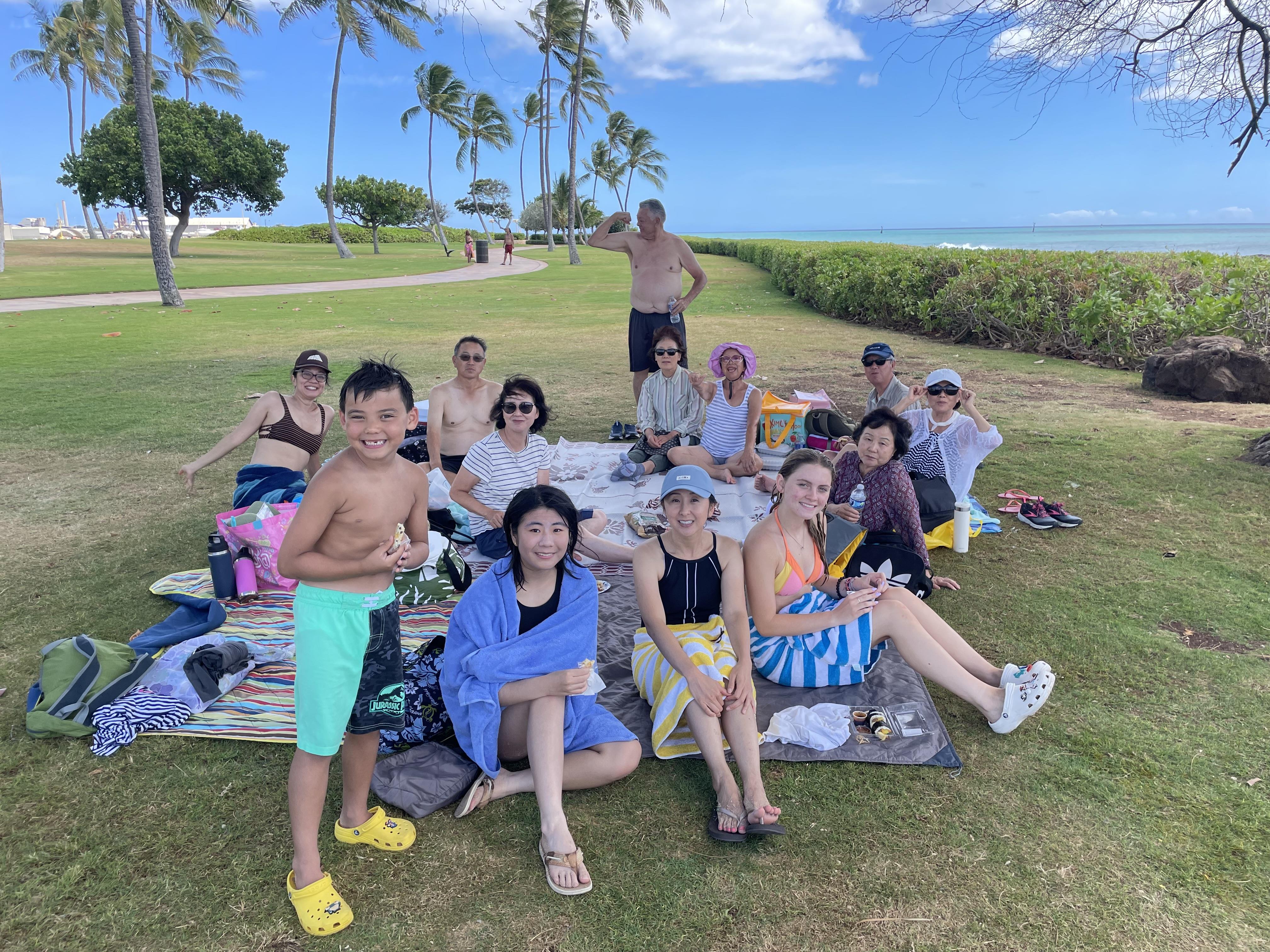 People gather for a picnic on grass with food and drinks under palm trees in Hawaii.