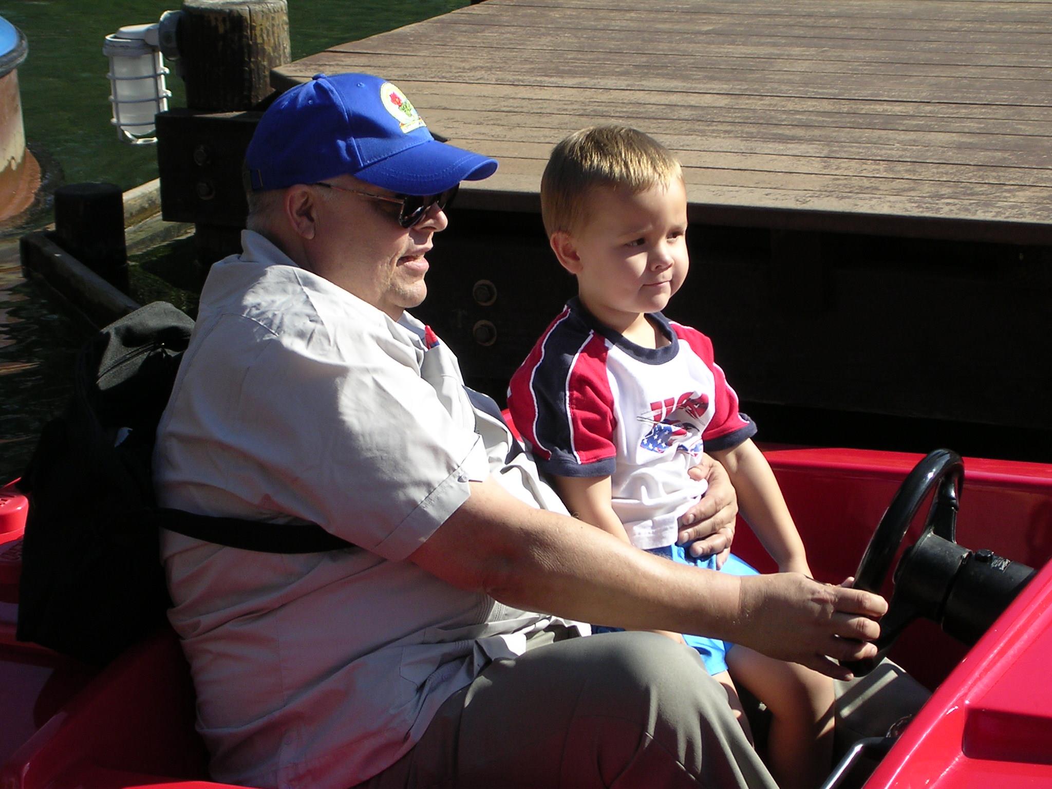 A man and a young boy are sitting on a boat. The man steers while the boy looks happy.