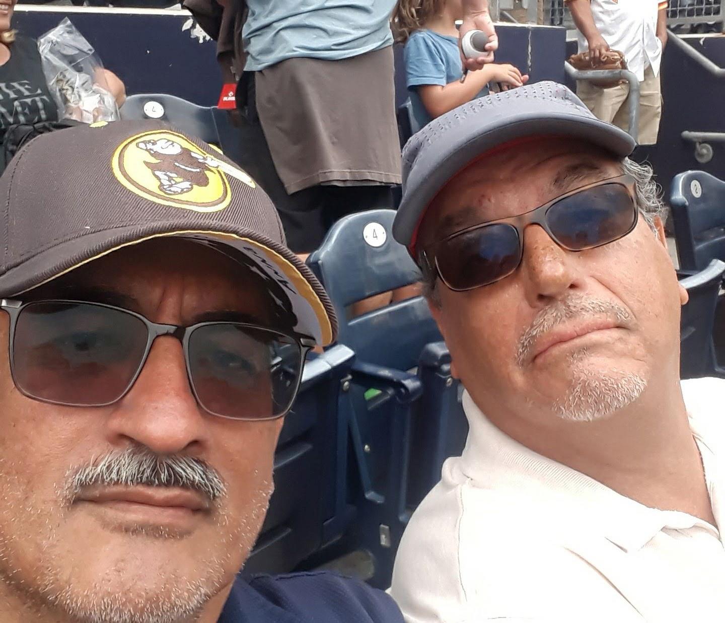Two men smile for the camera at a baseball game in the afternoon. They enjoy the moment together.