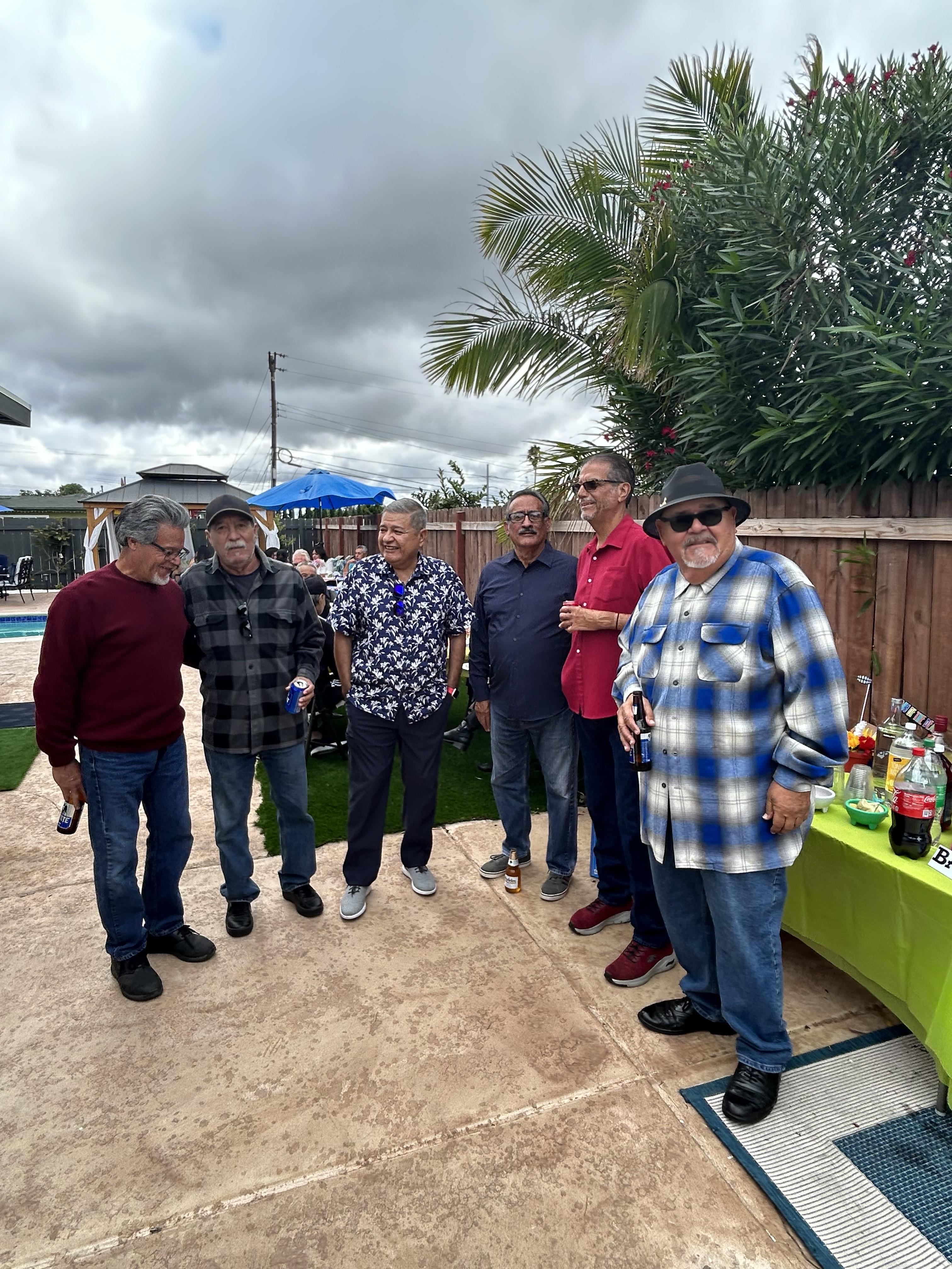 Six men stand together in a backyard during a party near a pool. The weather is cloudy.