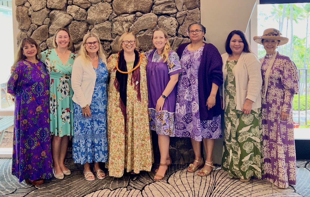 A group of women poses for a picture at a event beside a stone wall in the daytime.