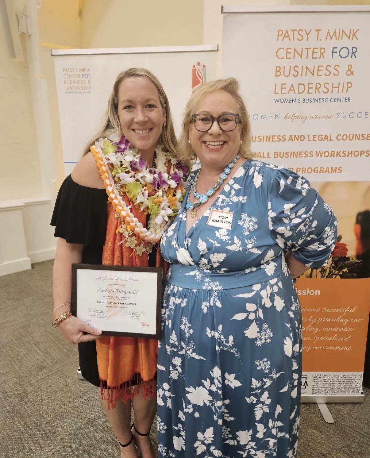 Two women hold a certificate at a business leadership event in a community center.