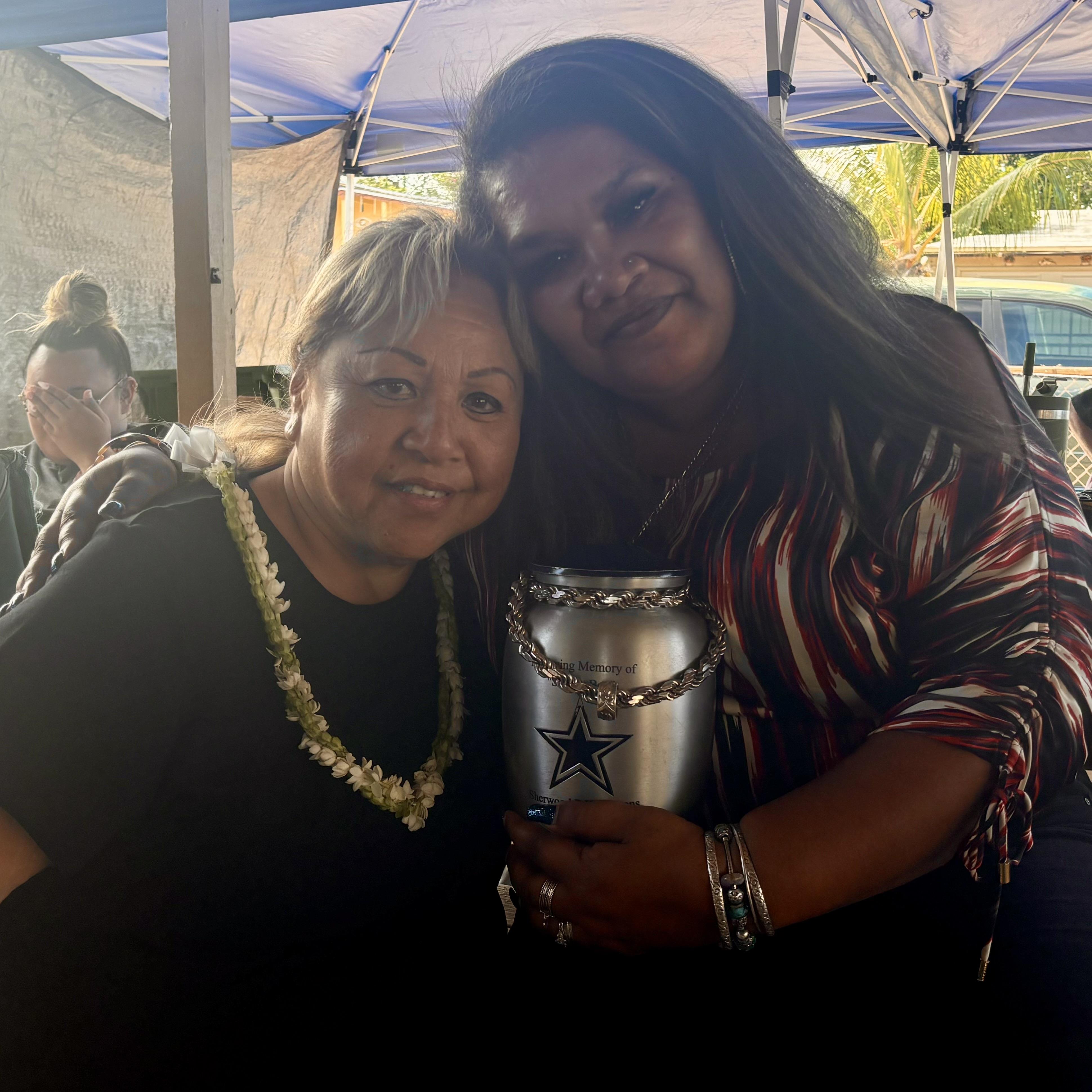 Two women stand close together, smiling while holding a silver container at a gathering outdoors.