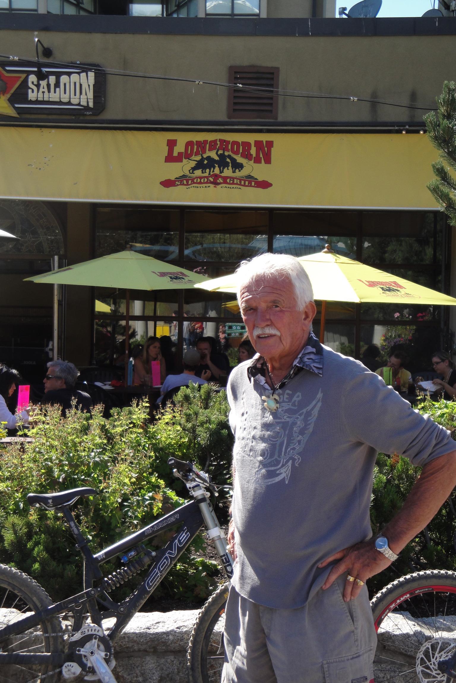 A man is near a restaurant with a bike. It is a bright day and people are dining outside.