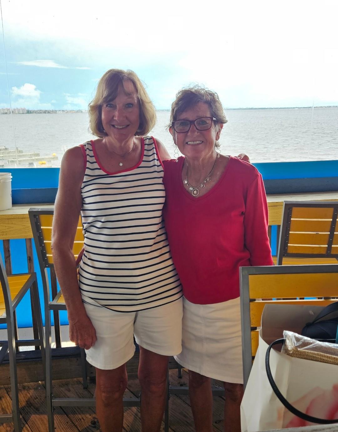 Two women stand side by side and smile at a beachside restaurant during daytime.