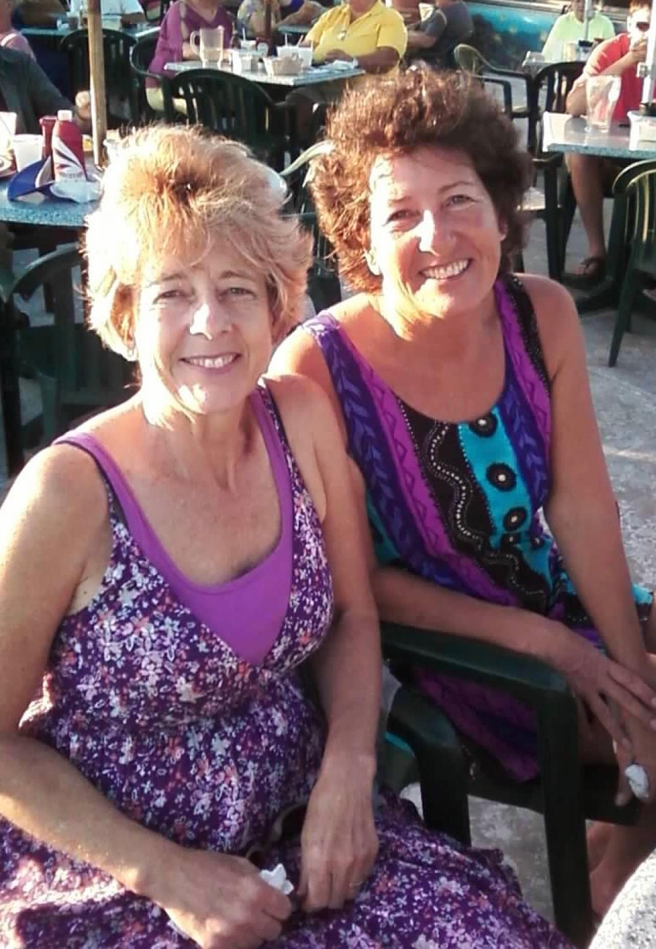 Two women smile as they sit together in outdoor seating during a sunny afternoon gathering.