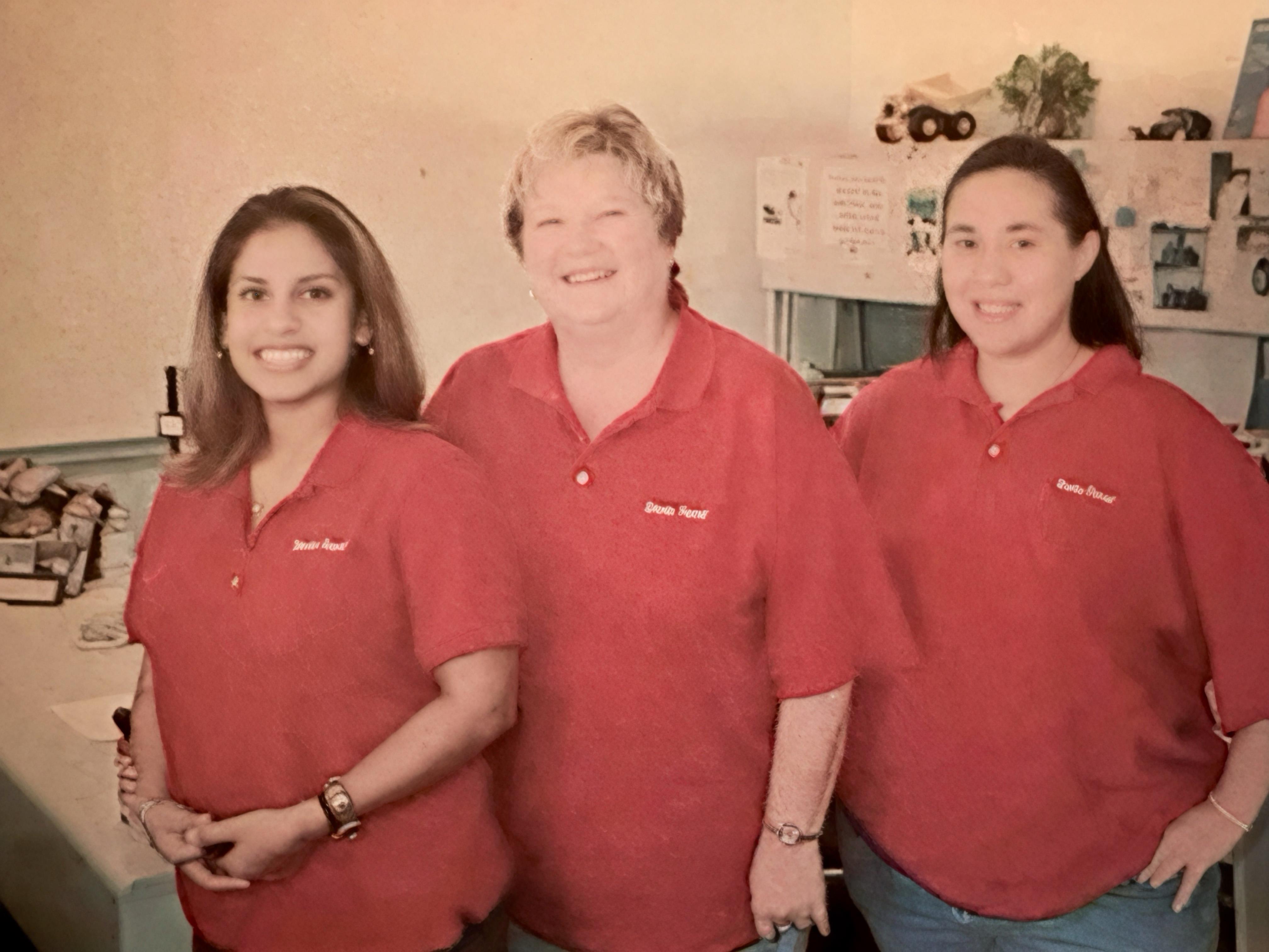 Three women smile and pose together in a work setting wearing matching red shirts.