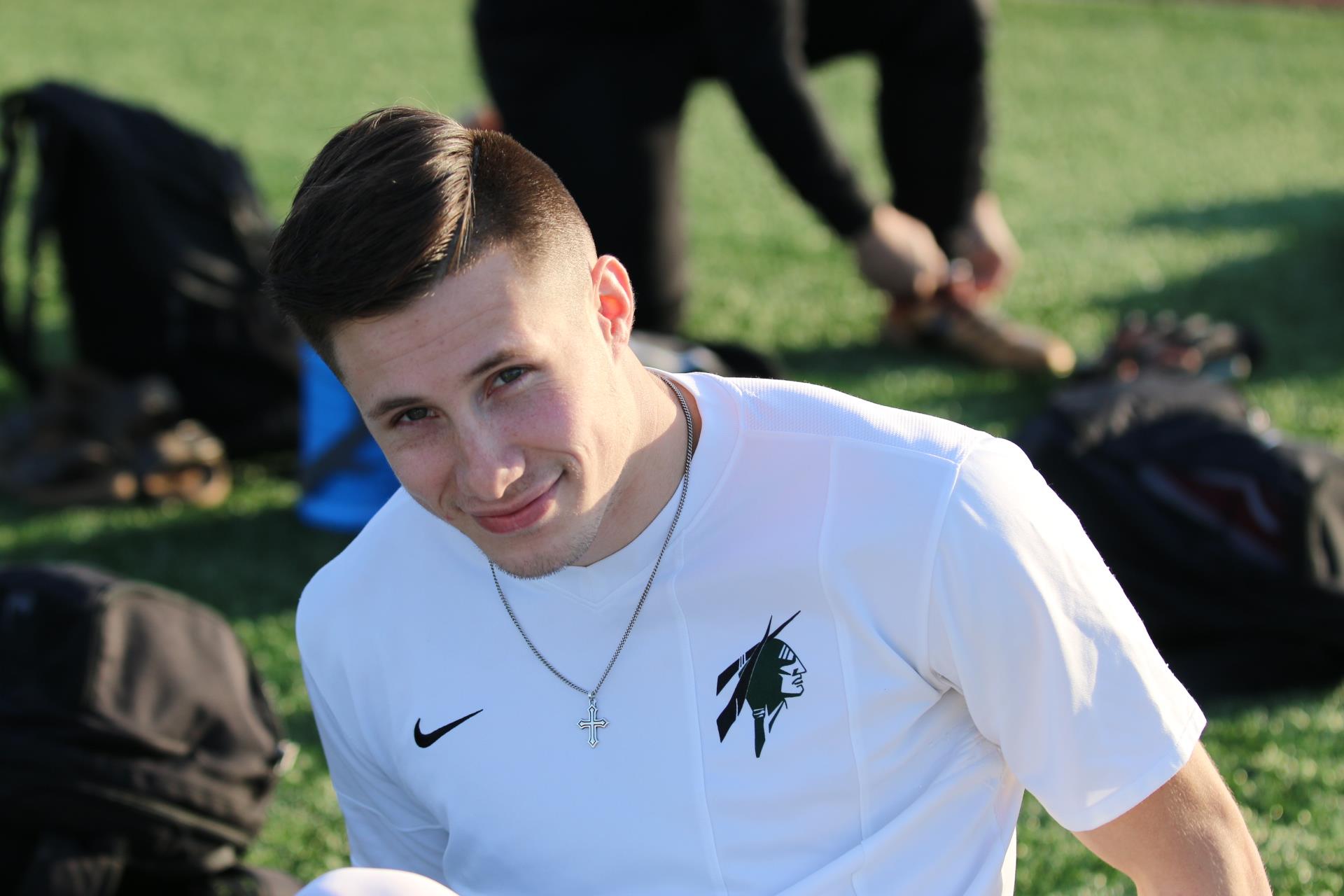 A young man sits on the grass at a sports field with others around him. He looks happy and engaged.