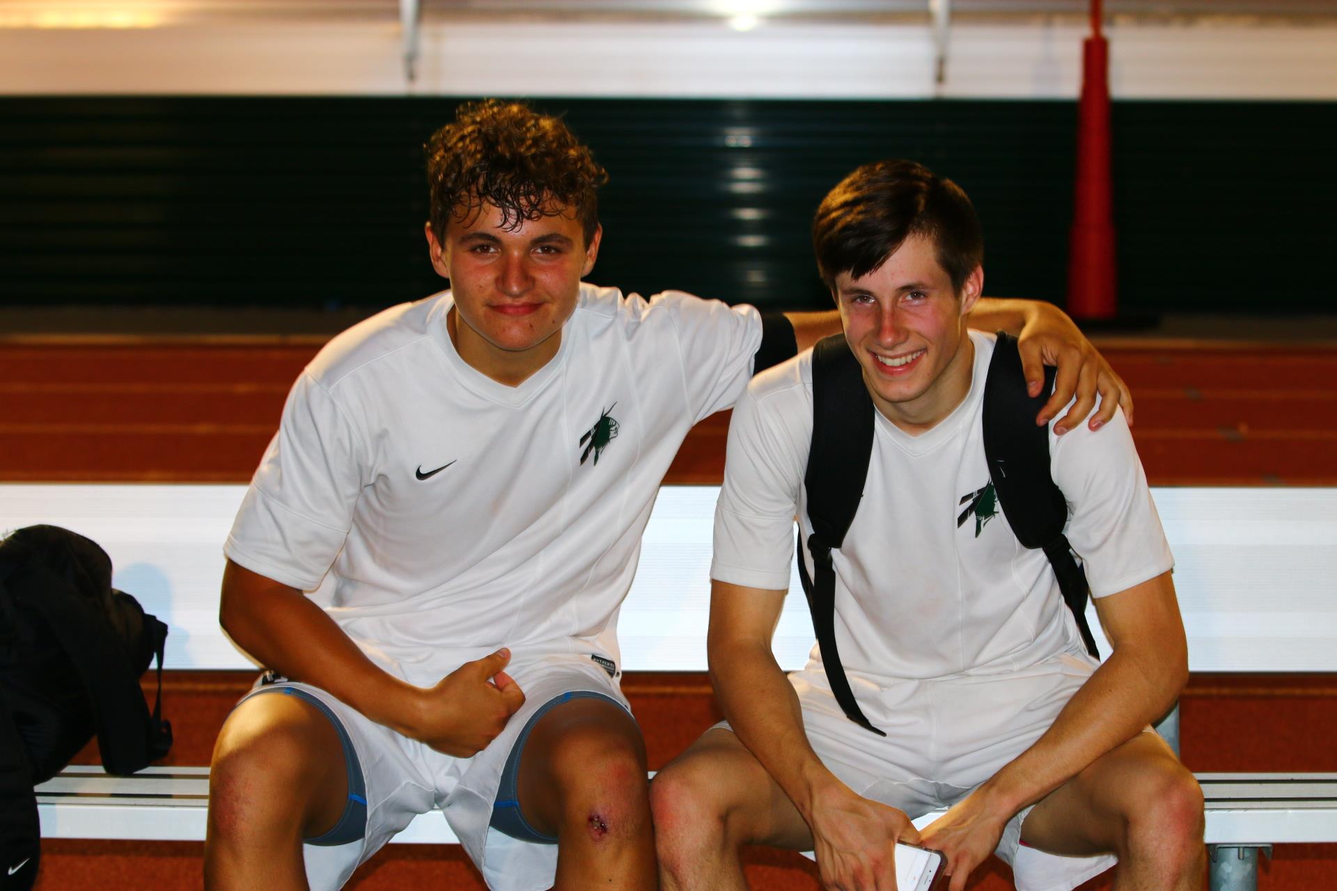Two boys wear white sports gear and sit on a bench, smiling, after playing soccer together.
