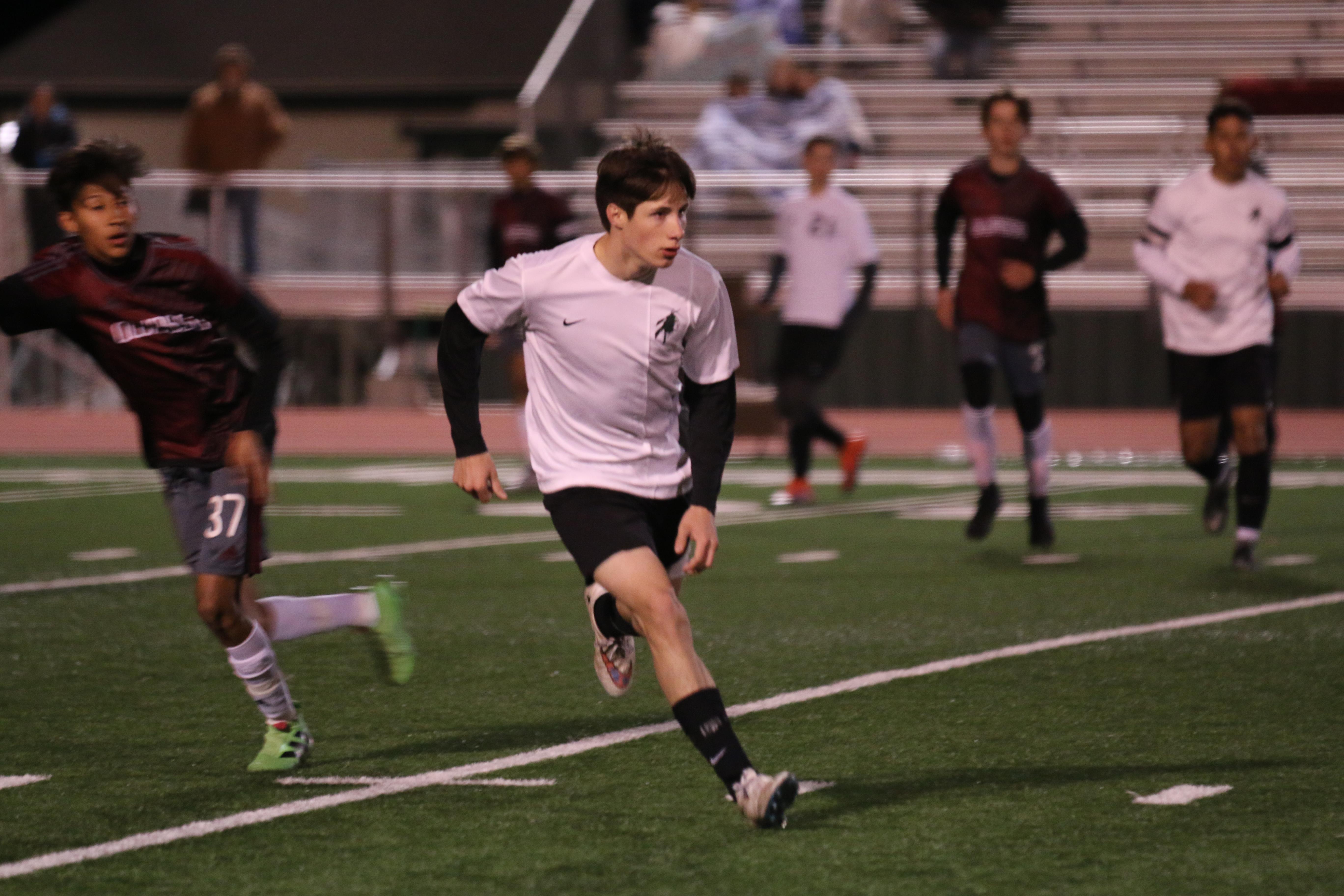 Players run on the soccer field during a night match with bright stadium lights shining down.