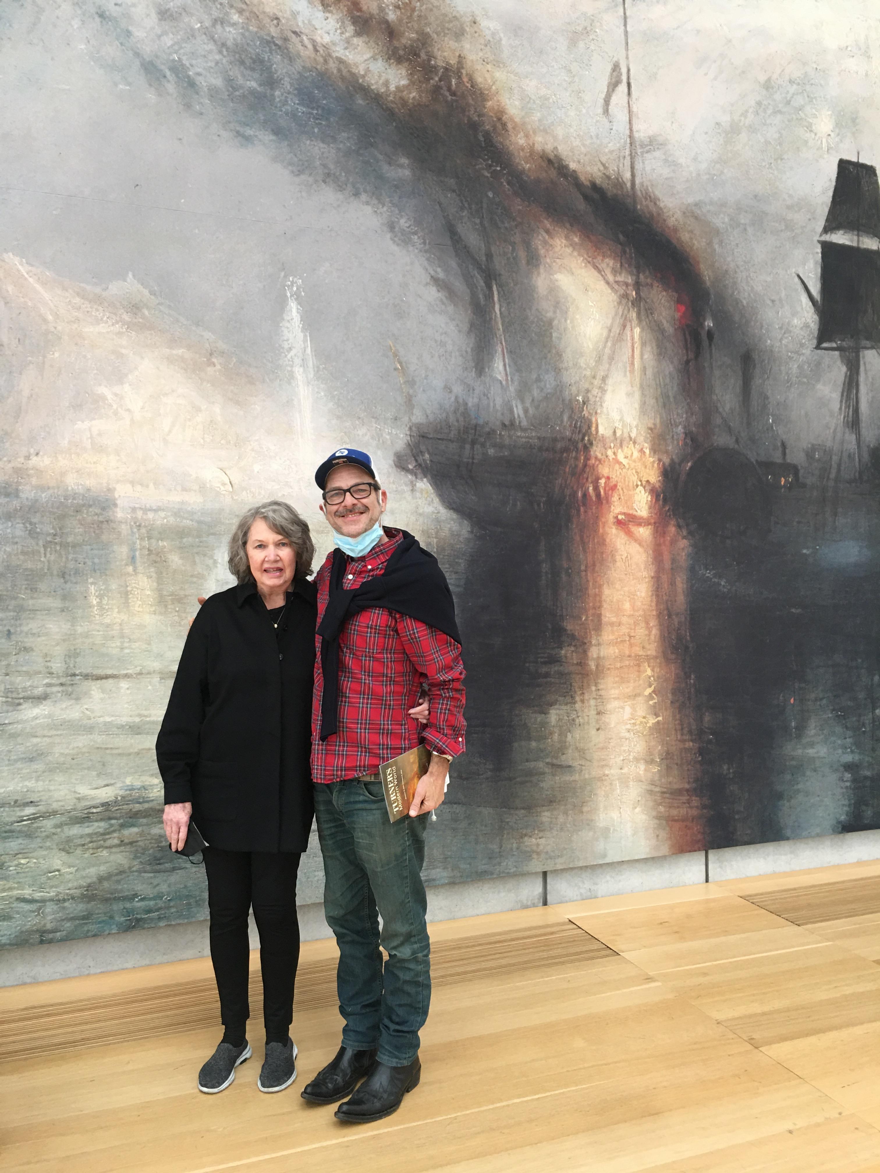 Two people pose in front of a mural showing a ship with smoke in an art gallery.