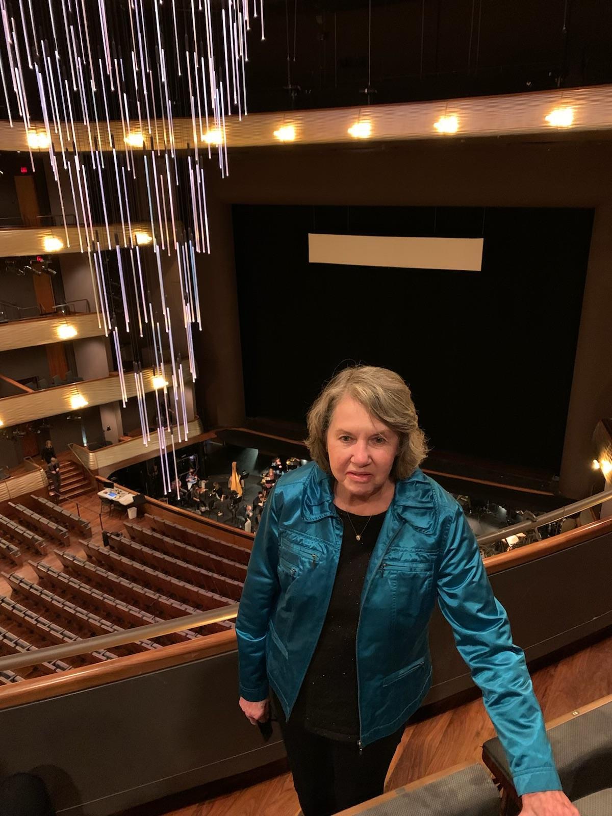 A woman poses near the edge of a balcony in a theater. Actors prepare below on the stage.