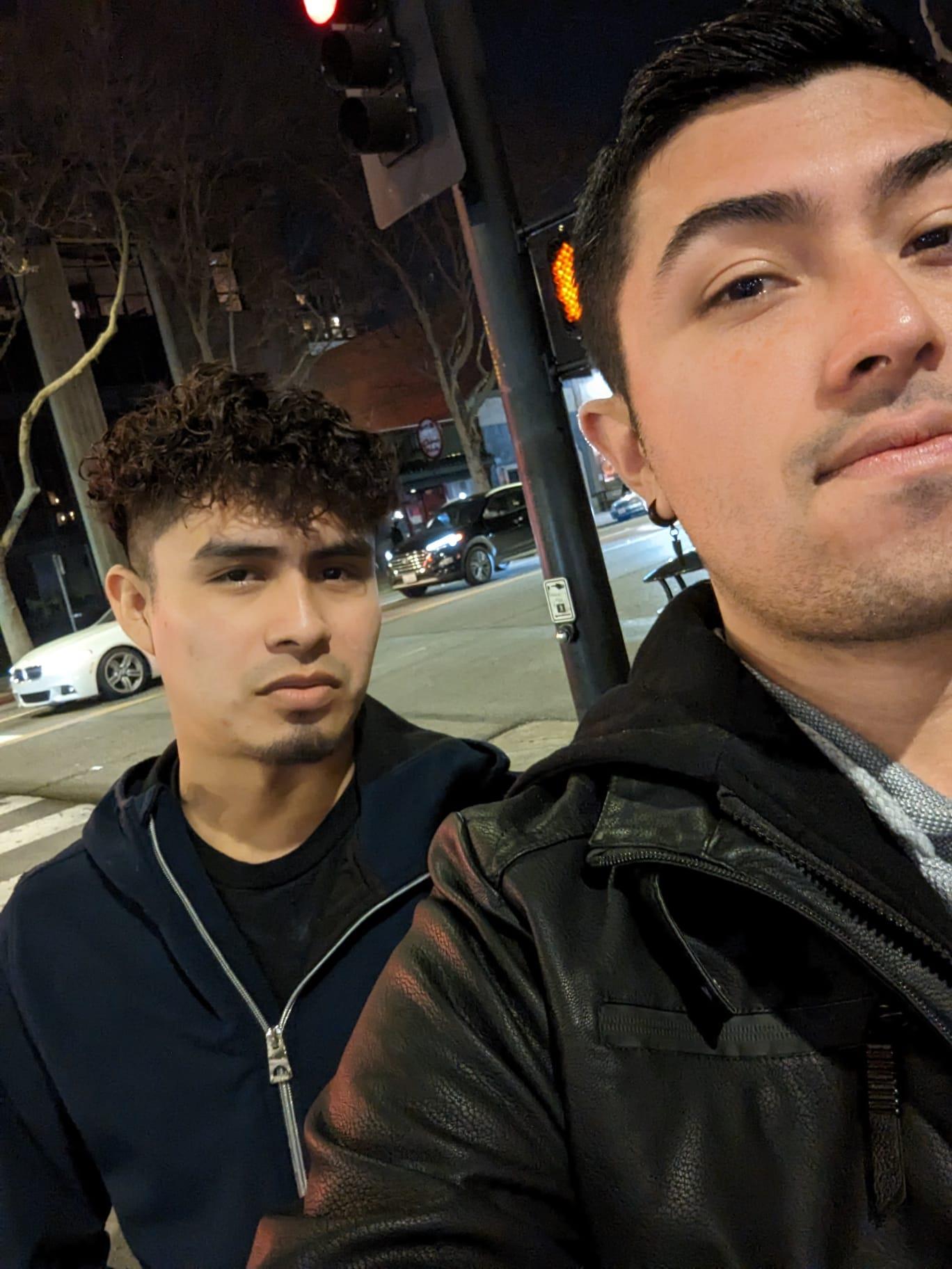 Two young men are outside on a busy street at night with cars and lights around them.