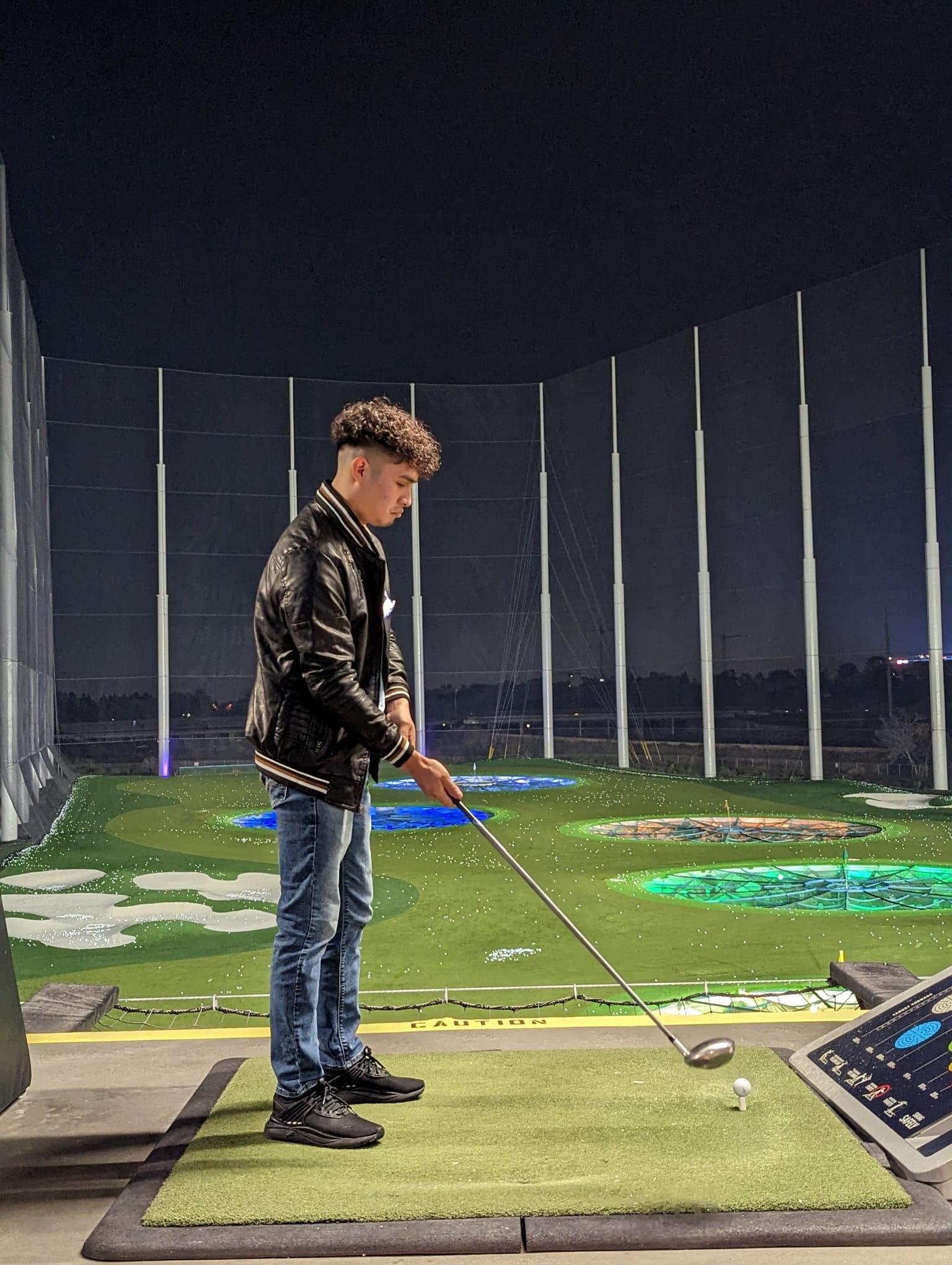 A young man stands on a golf tee preparing to swing at a driving range at night.