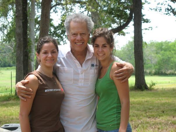 Three people are smiling and posing together in a park setting during the day among green trees.