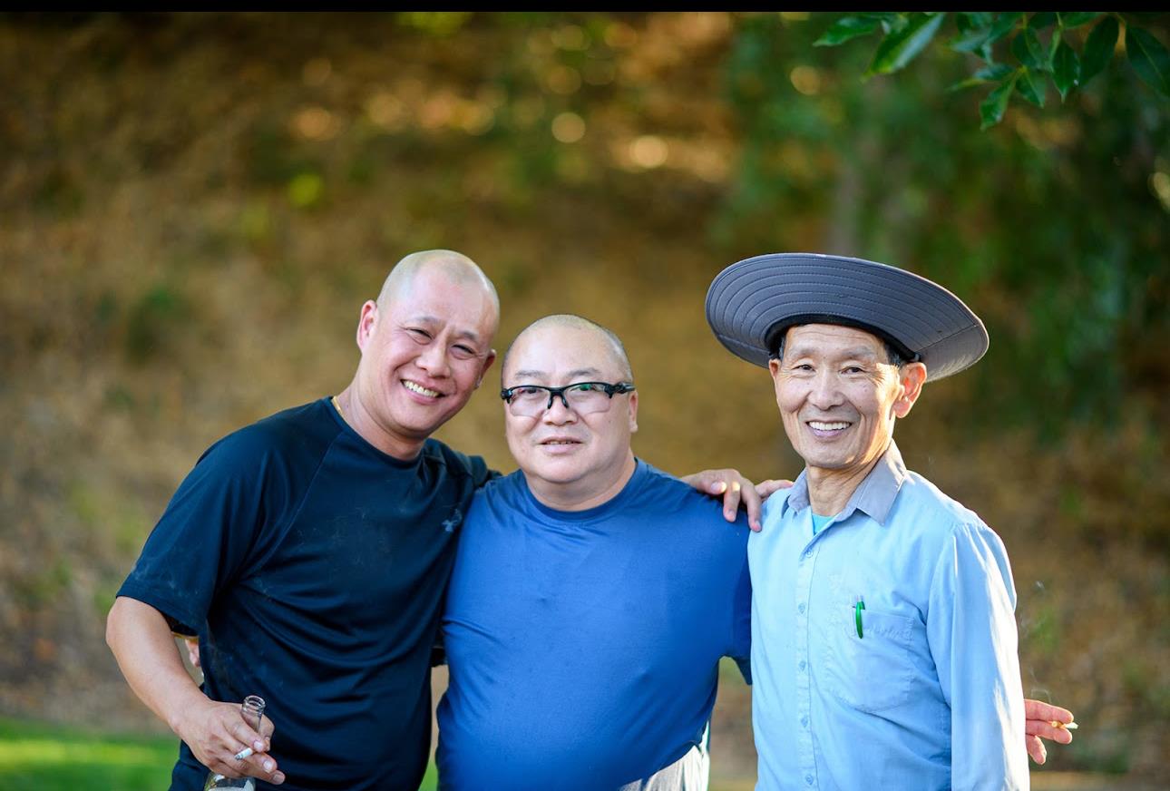 Three men stand close together smiling outdoors on a sunny day in a natural setting.
