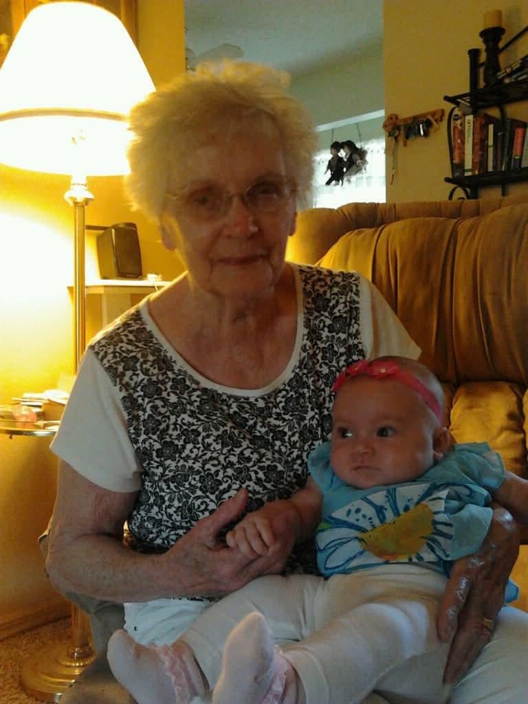 An older woman sits in a chair while holding a baby in her lap. The room has warm lights.