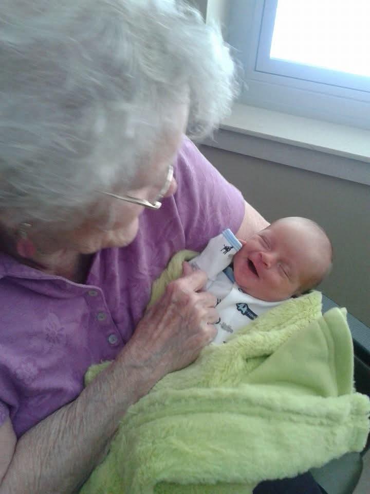An elderly woman smiles in a chair by the window, holding a baby boy in a green blanket.
