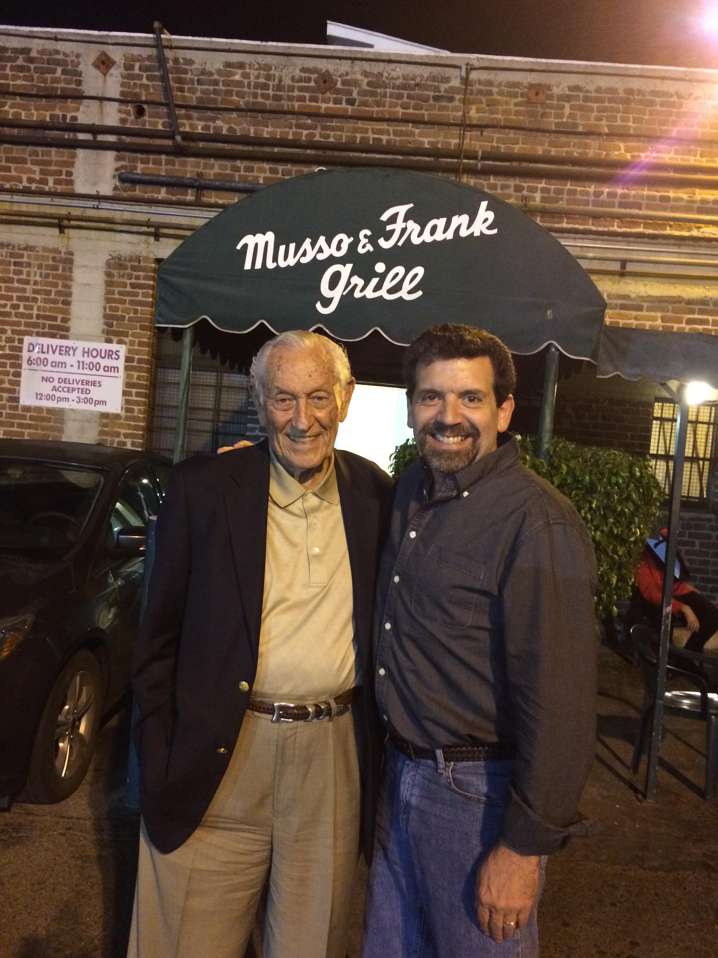 Two men smile and pose for a picture outside Musso and Frank Grill in Los Angeles at night.