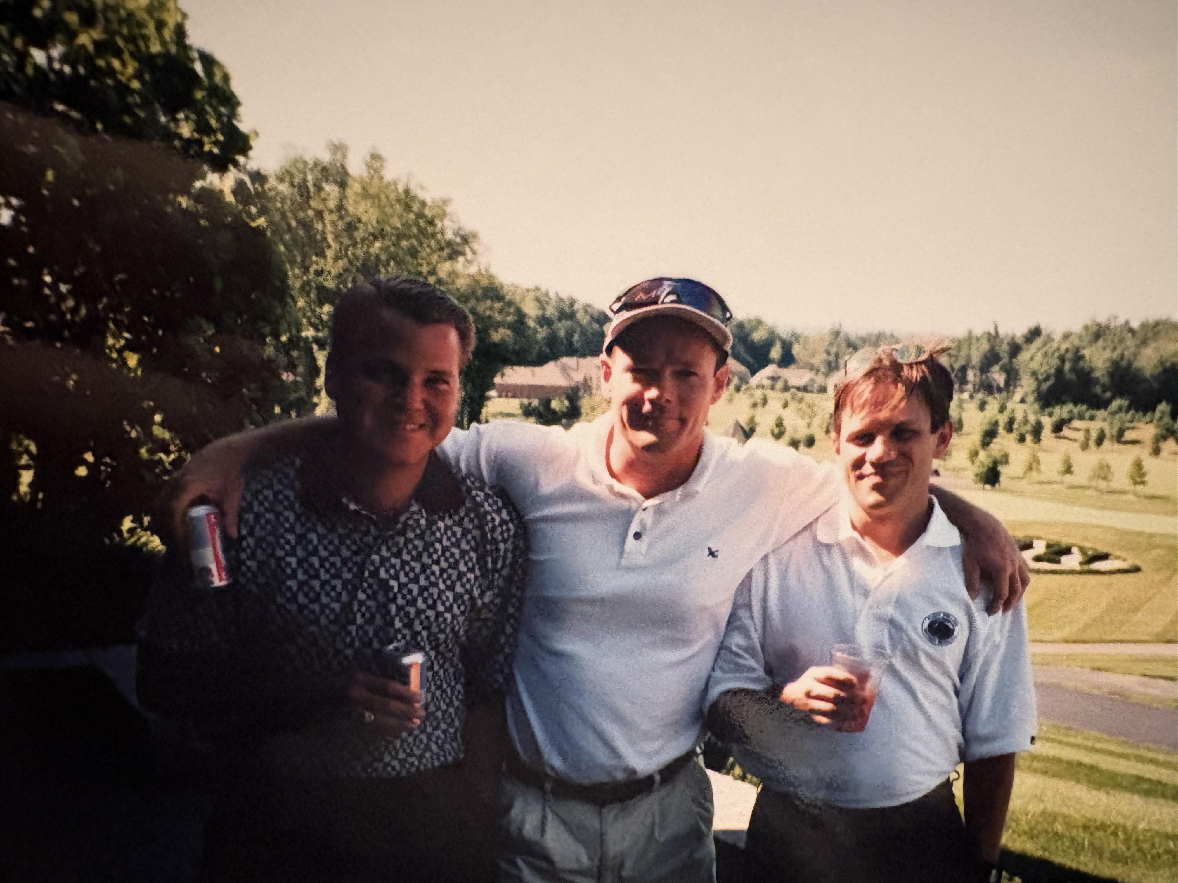Three friends stand close together while holding drinks and smiling on the golf course.