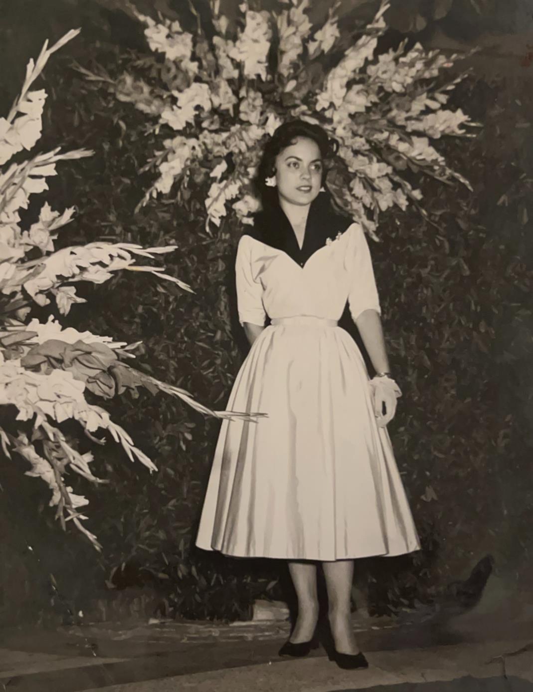 A woman stands beside large flowers at a formal event, wearing an elegant outfit and smiling.