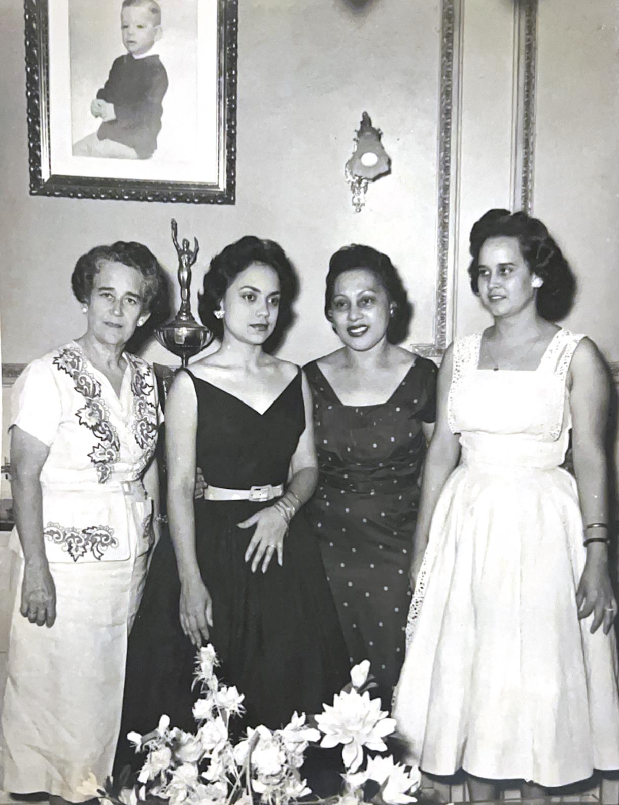 Four women stand together in a room, dressed in varying styles of dresses, smiling at the camera.