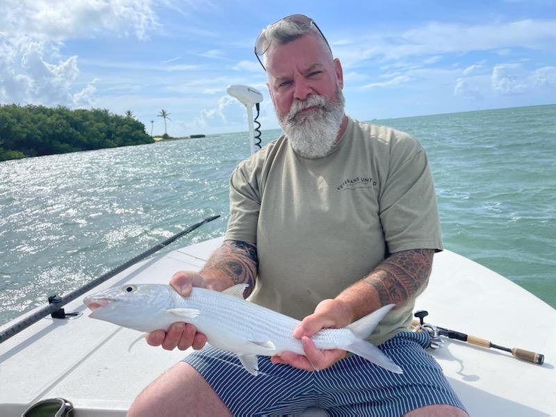 A man holds a fish on a boat in the water. The sky is clear with some clouds.