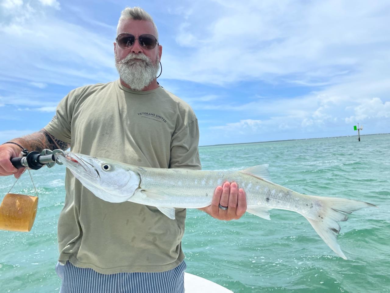 A man holds a large fish while standing on a boat in clear blue water. It is daytime and sunny.