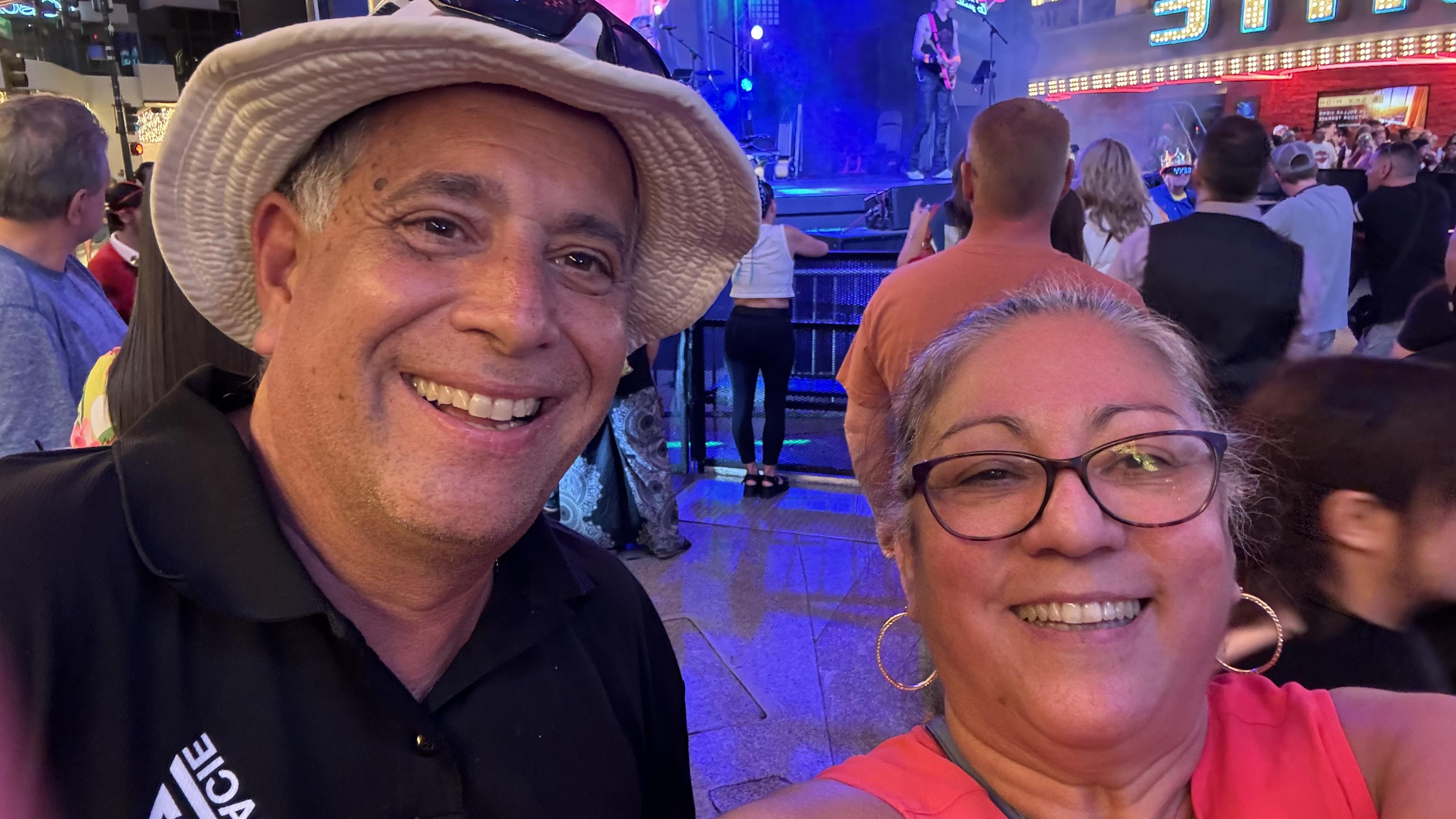 Couple smiles widely while attending an outdoor concert with colorful lights in the background.