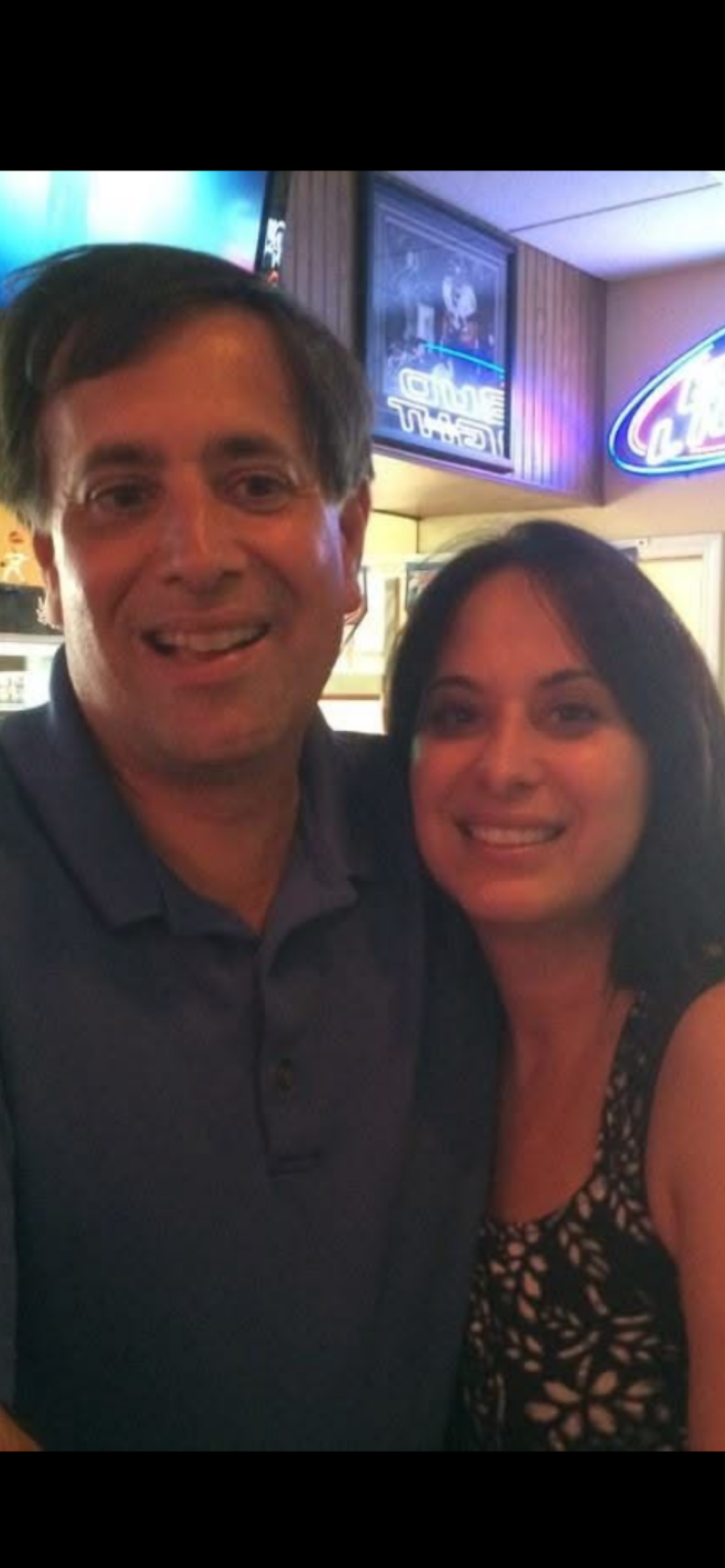 Two people are smiling while standing close together in a social setting, enjoying their time.
