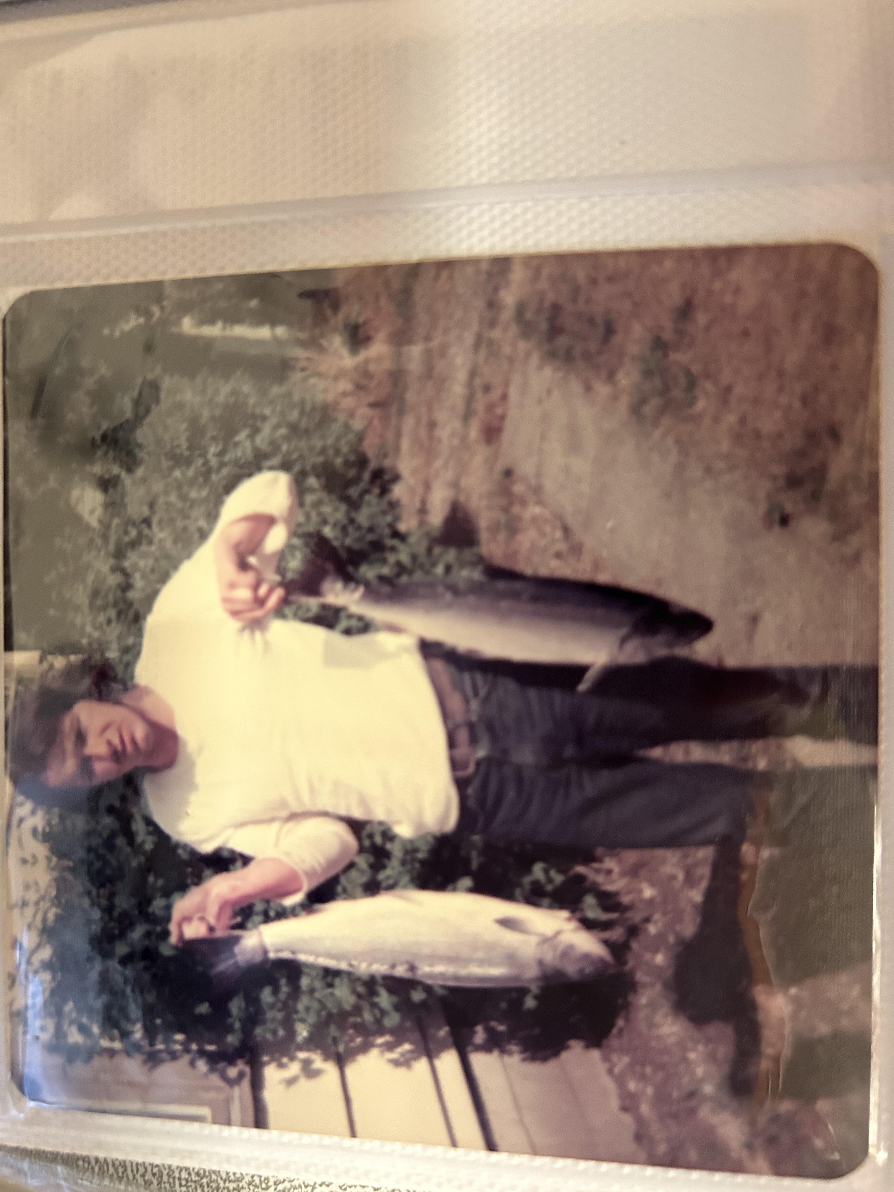 A man stands near a lake with two large fish, smiling and posing for the camera in the sun.