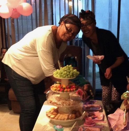 Two friends are standing near a table filled with snacks and fruits during a gathering.