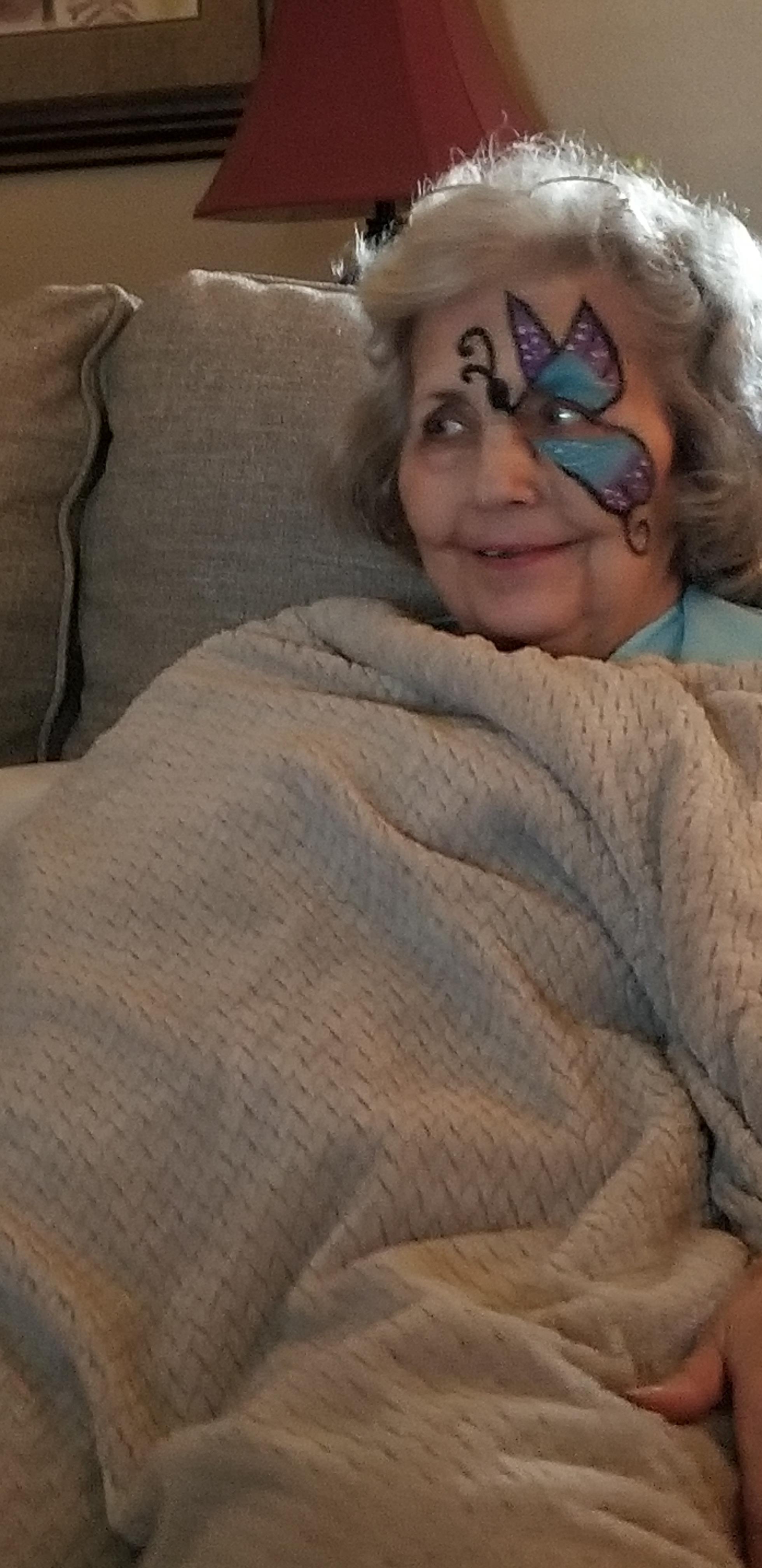 An elderly woman sits on a couch with a blanket while showing a face design and smiling.