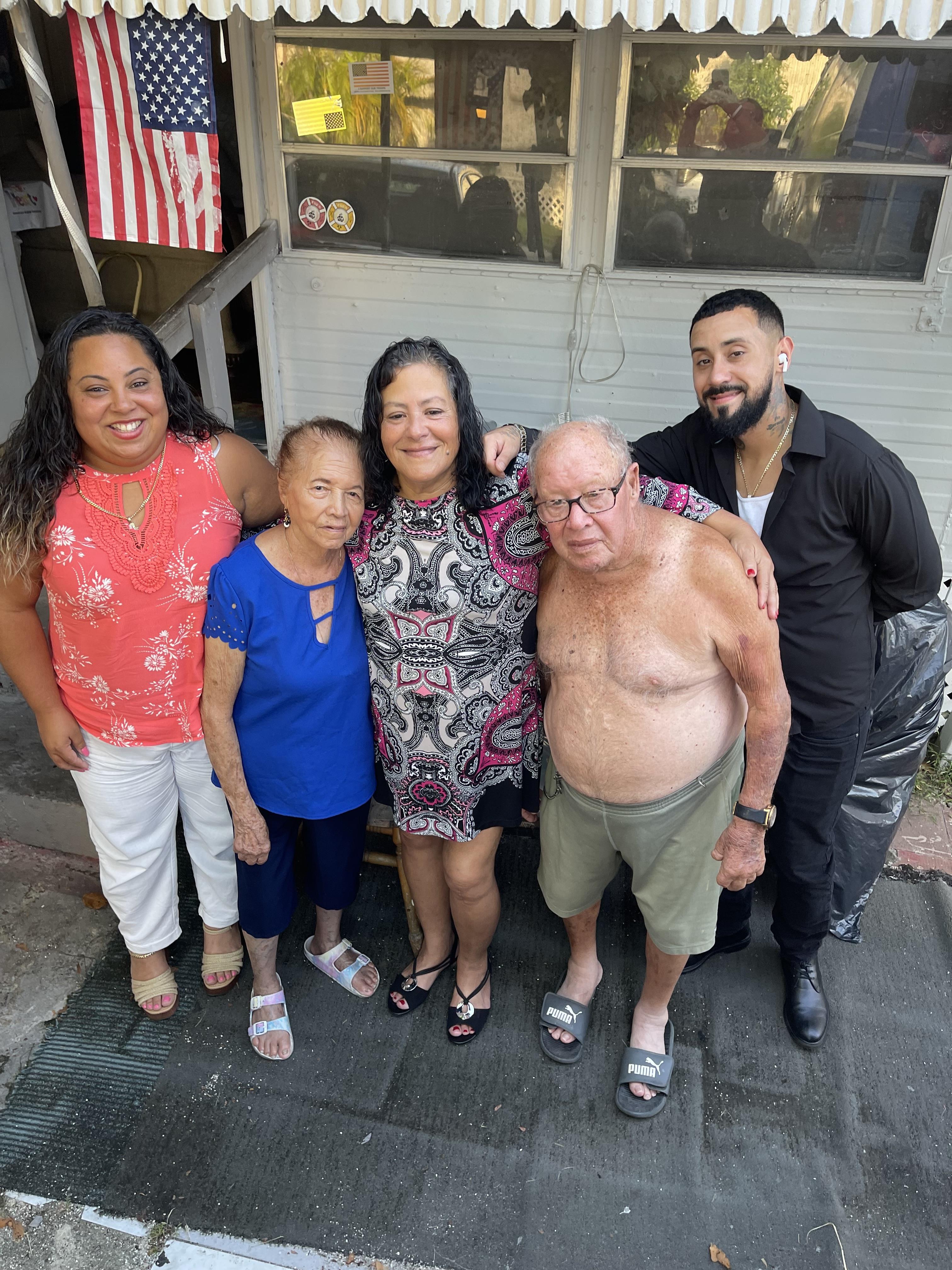 Family members stand together outside their house smiling and posing for the camera in the sun.