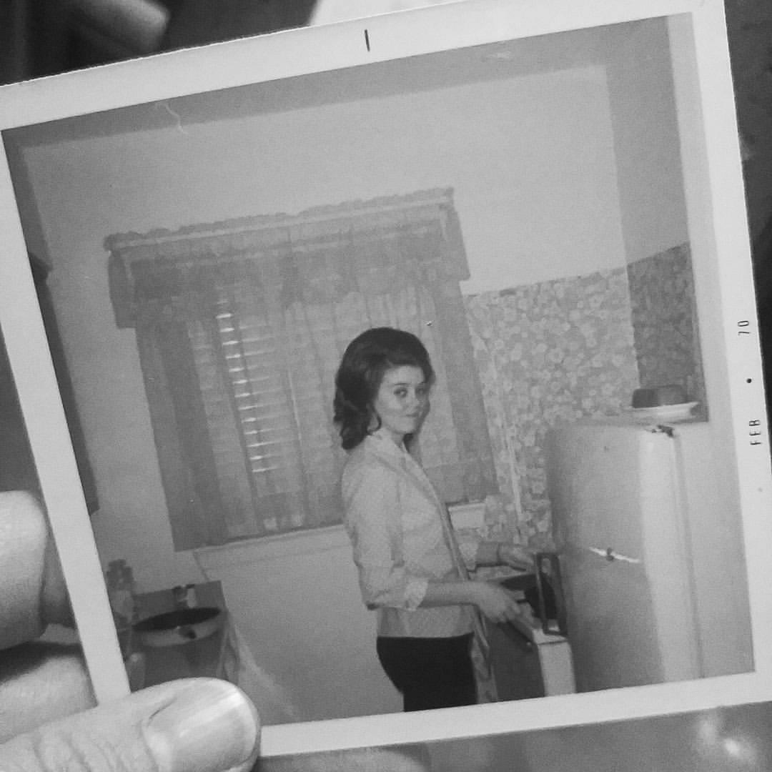 A hand holding a polaroid of a woman in a kitchen