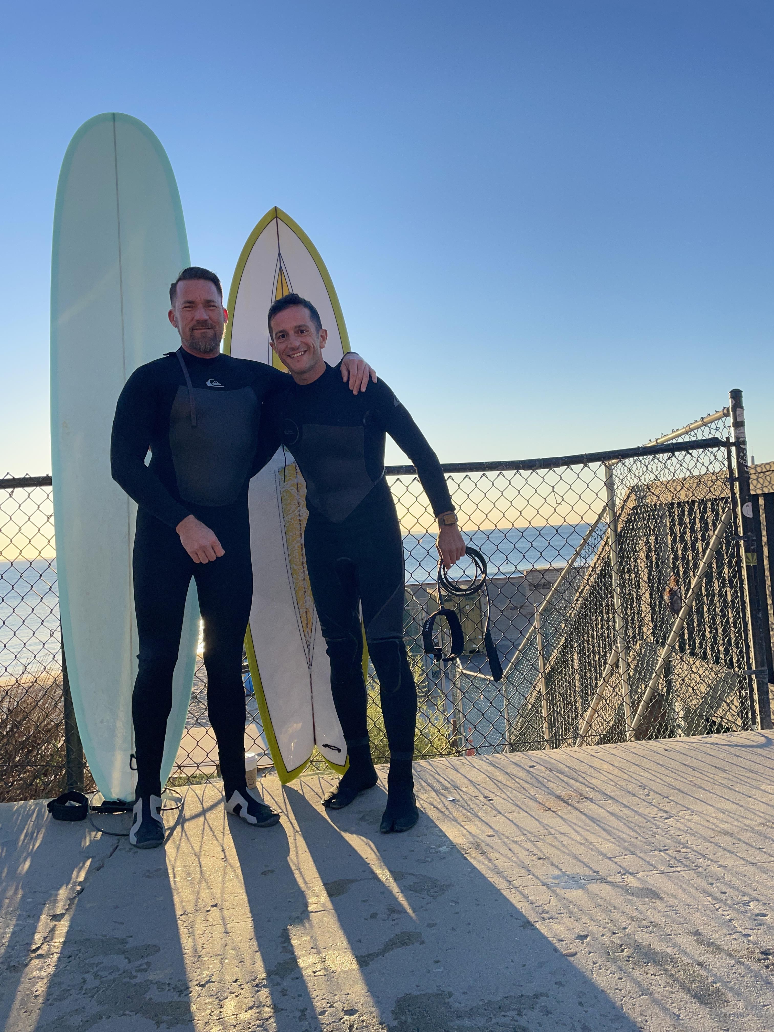 Two men in wet suits standing next to a surfboard