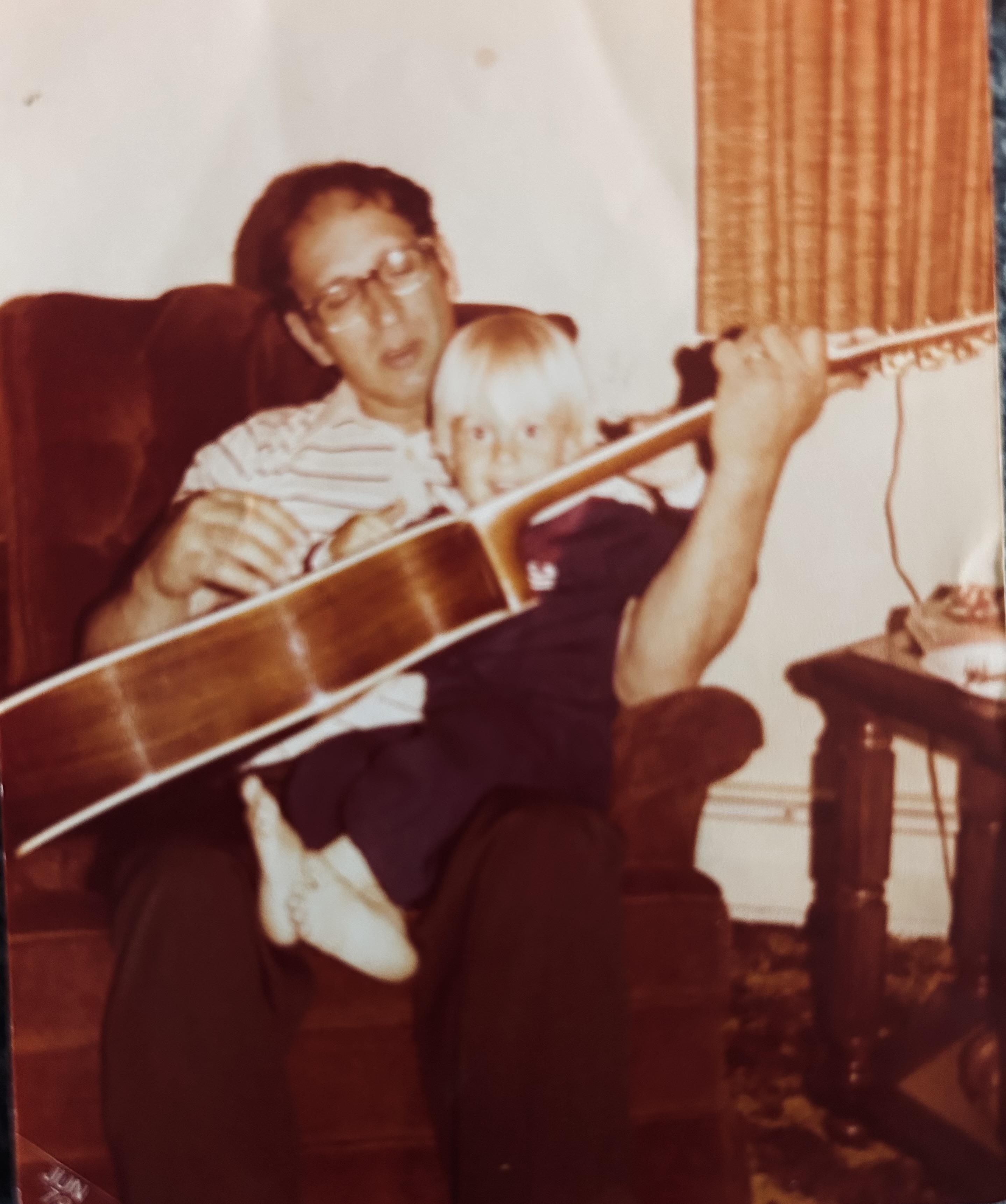 A man and child sitting in a chair playing guitar