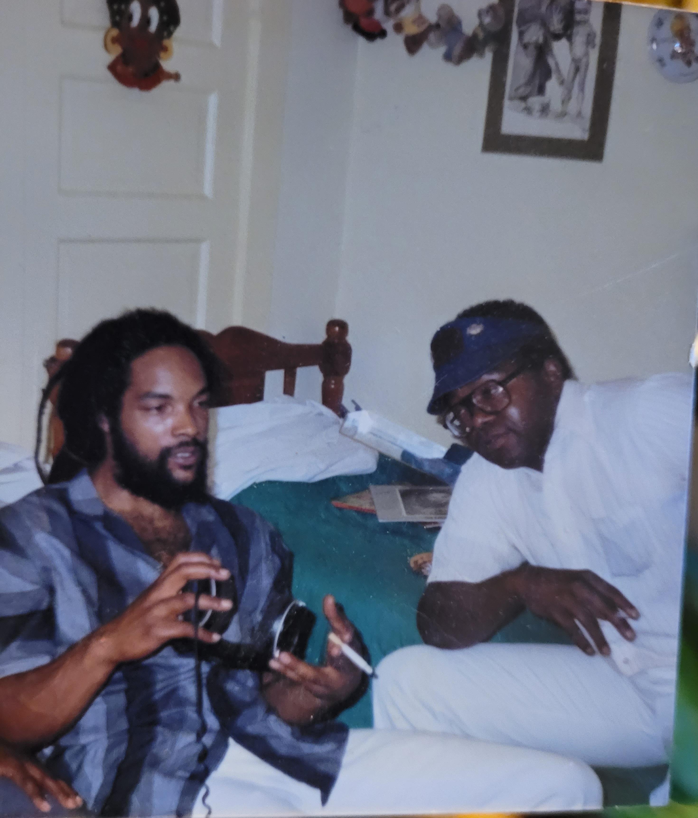 A man holding a camera and another man sitting on a bed