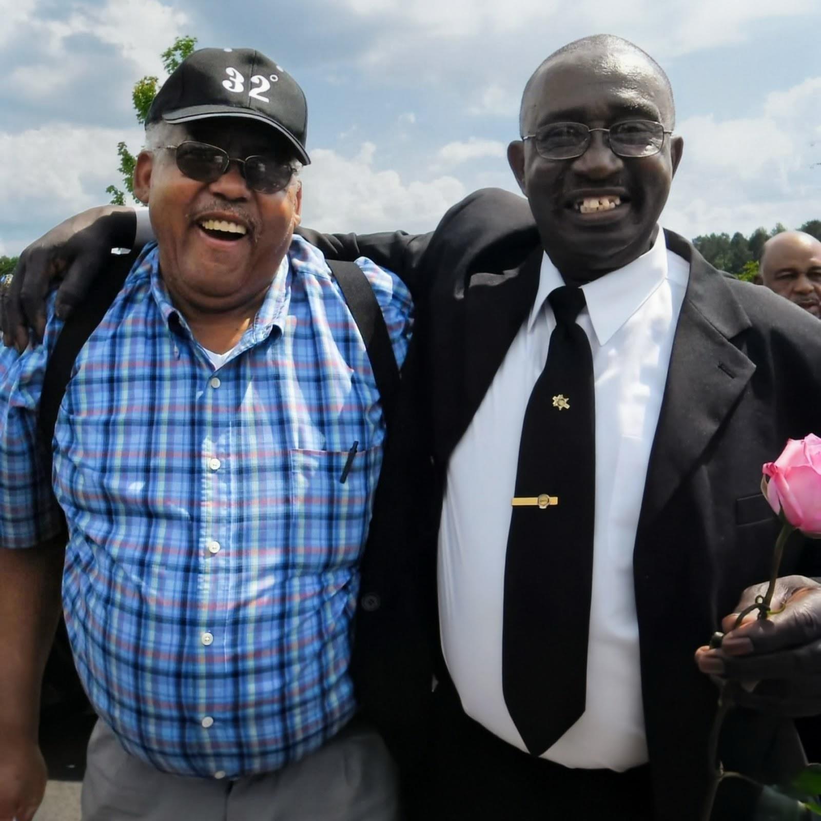 A couple of men smiling and holding a rose