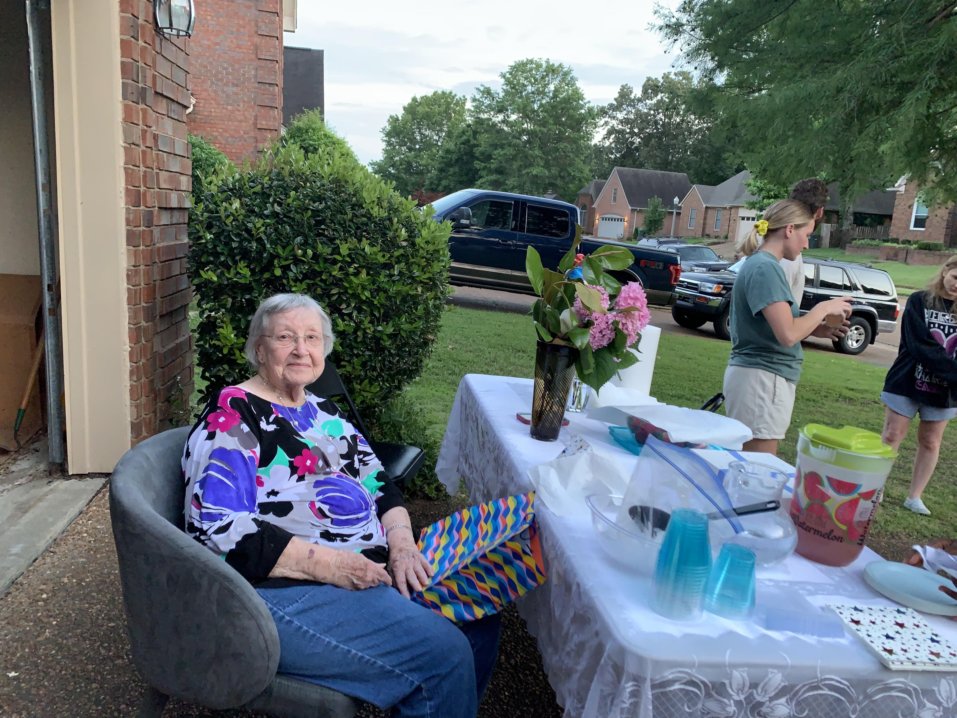 An older woman sitting in a chair outside with a table with flowers and a woman standing behind her