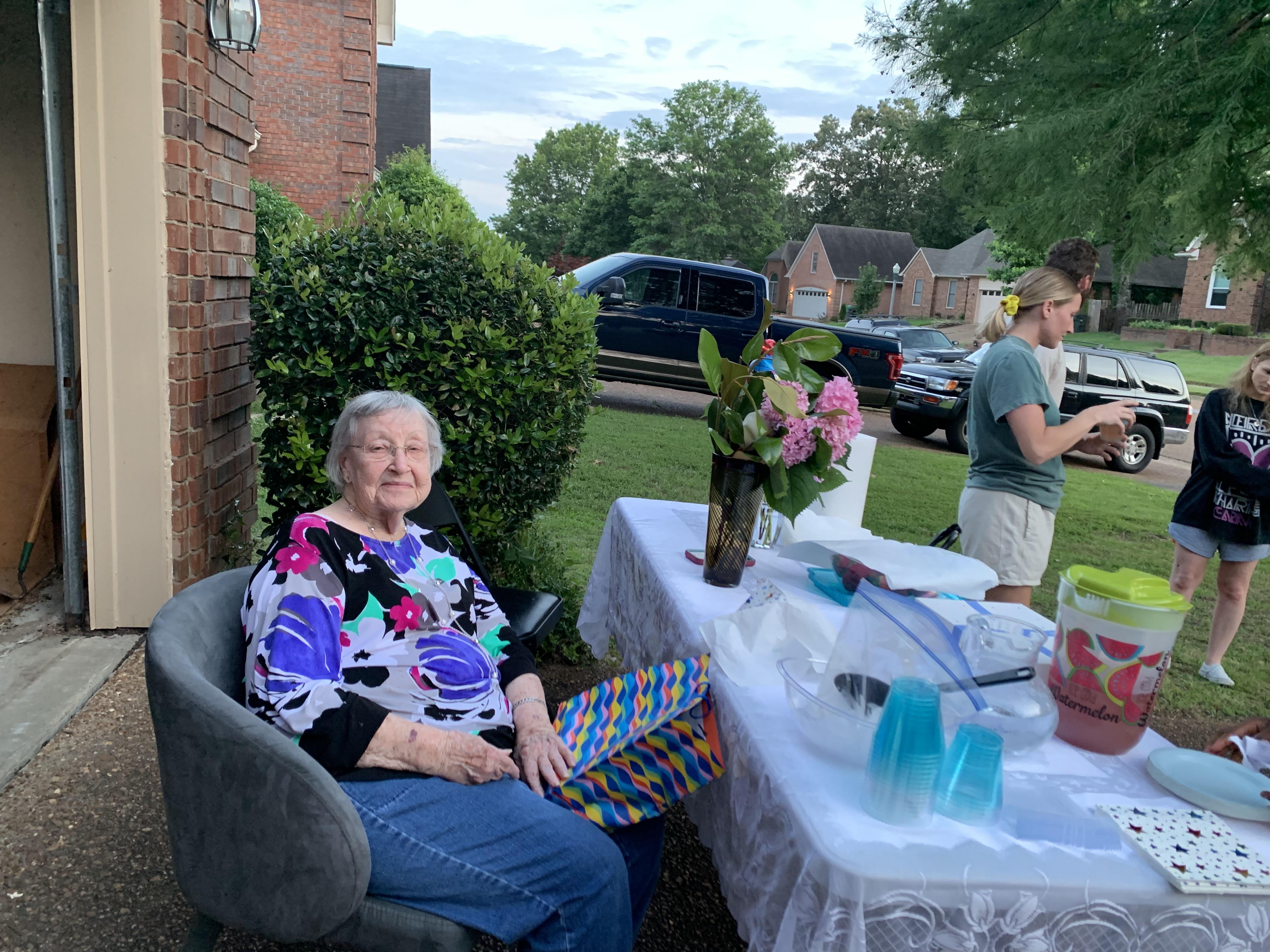 An old woman sitting in a chair outside with a table with flowers and a woman standing behind her