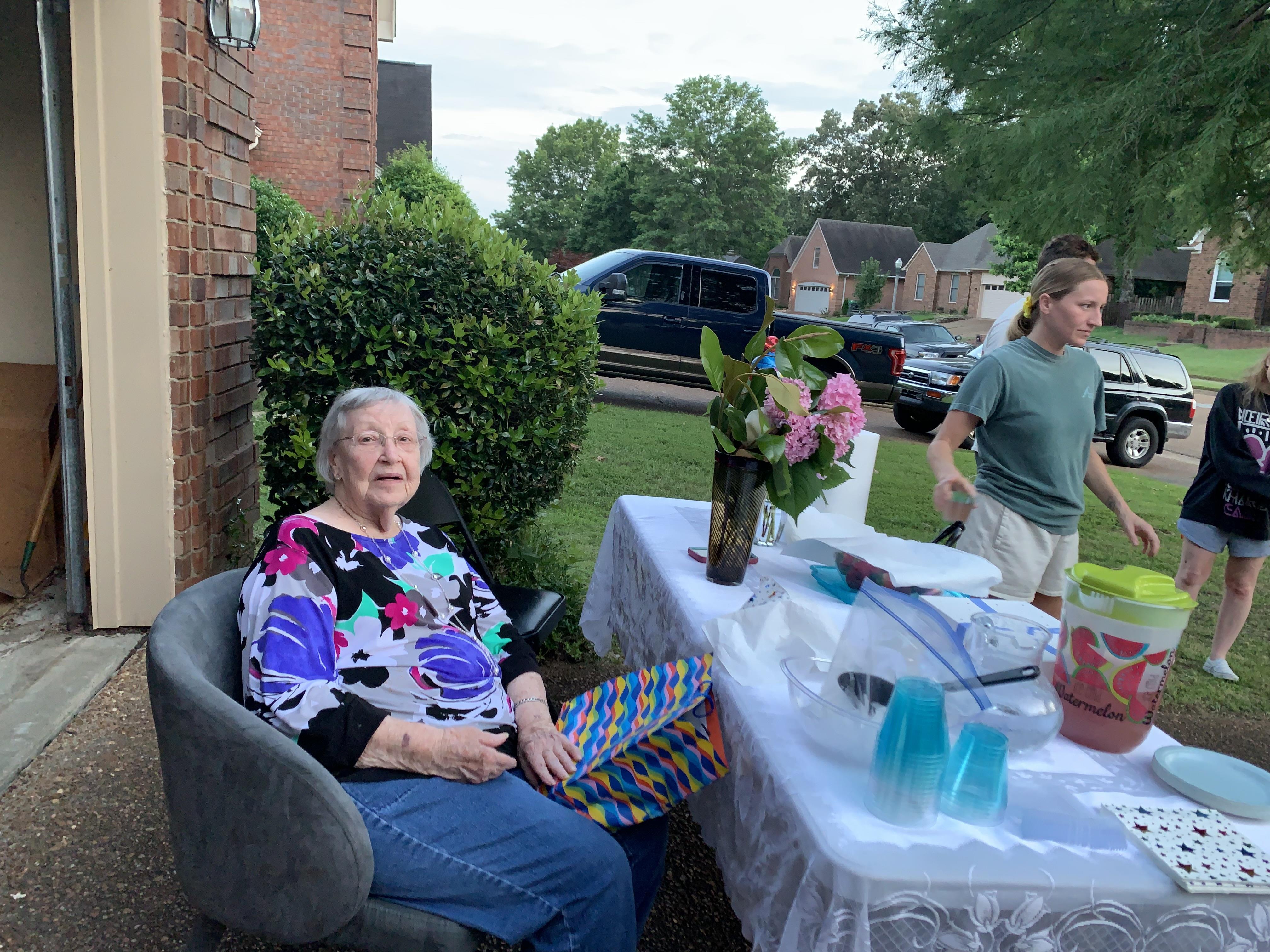 An older woman sitting in a chair outside with a table with flowers and a man standing behind her