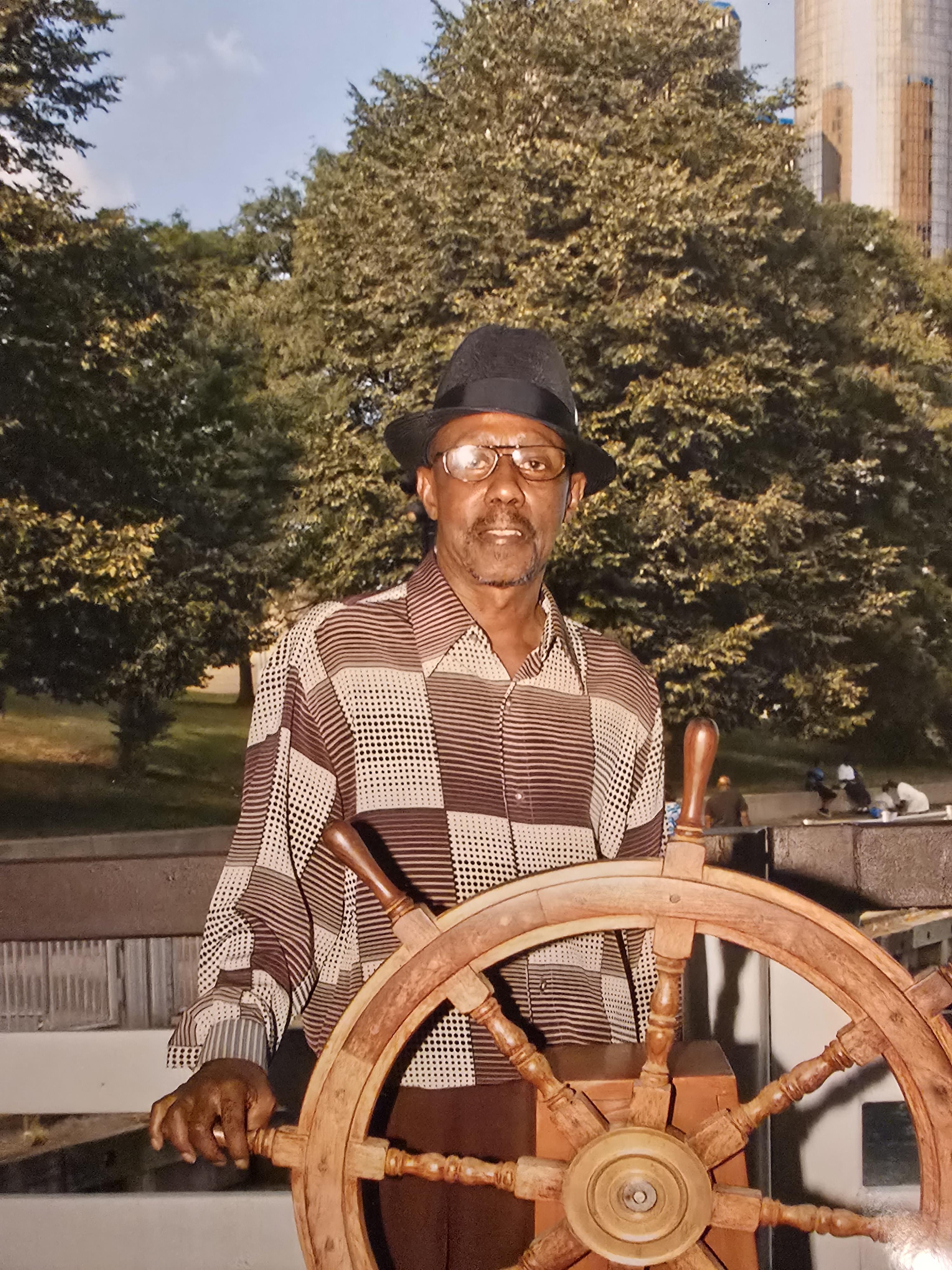A man in a hat and glasses holding a wooden steering wheel