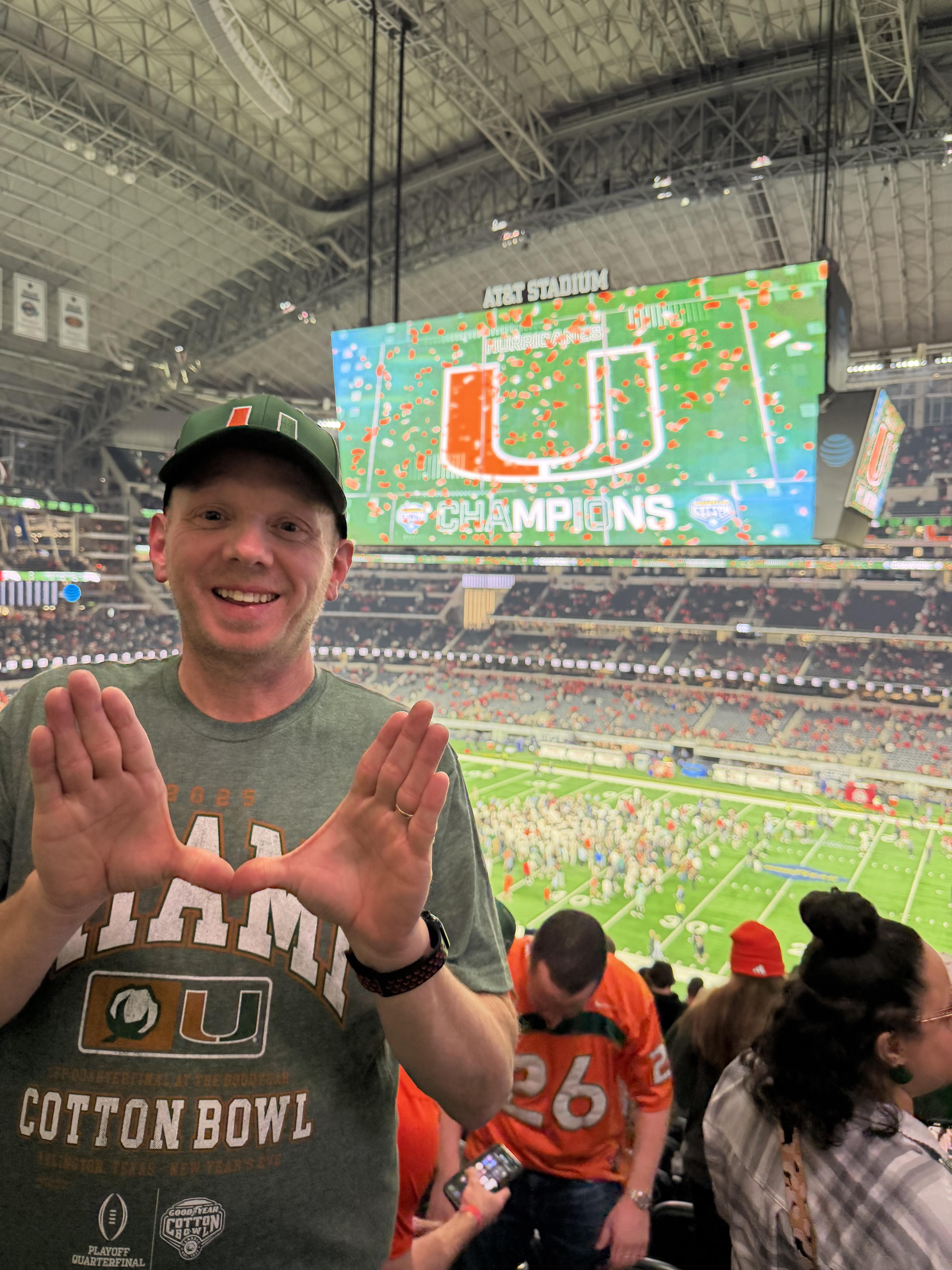 A man in a green hat with his hands up in front of a large stadium
