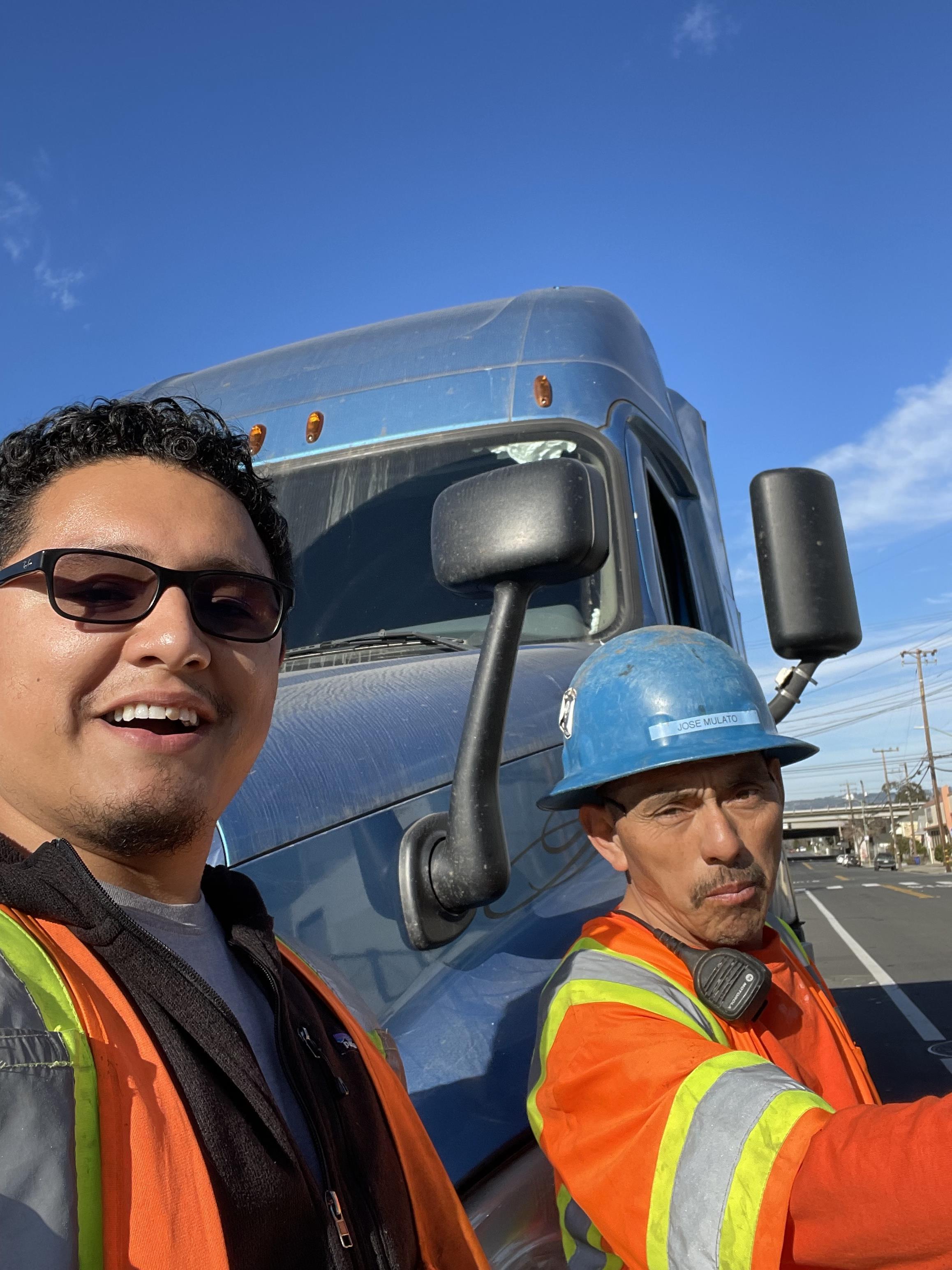 A selfie of two men in safety vests and a blue hard hat