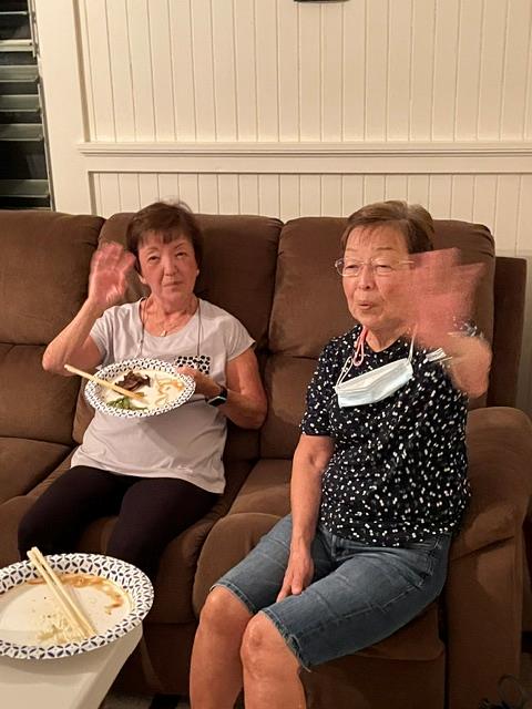 Two women sitting on a couch with plates of food and a mask