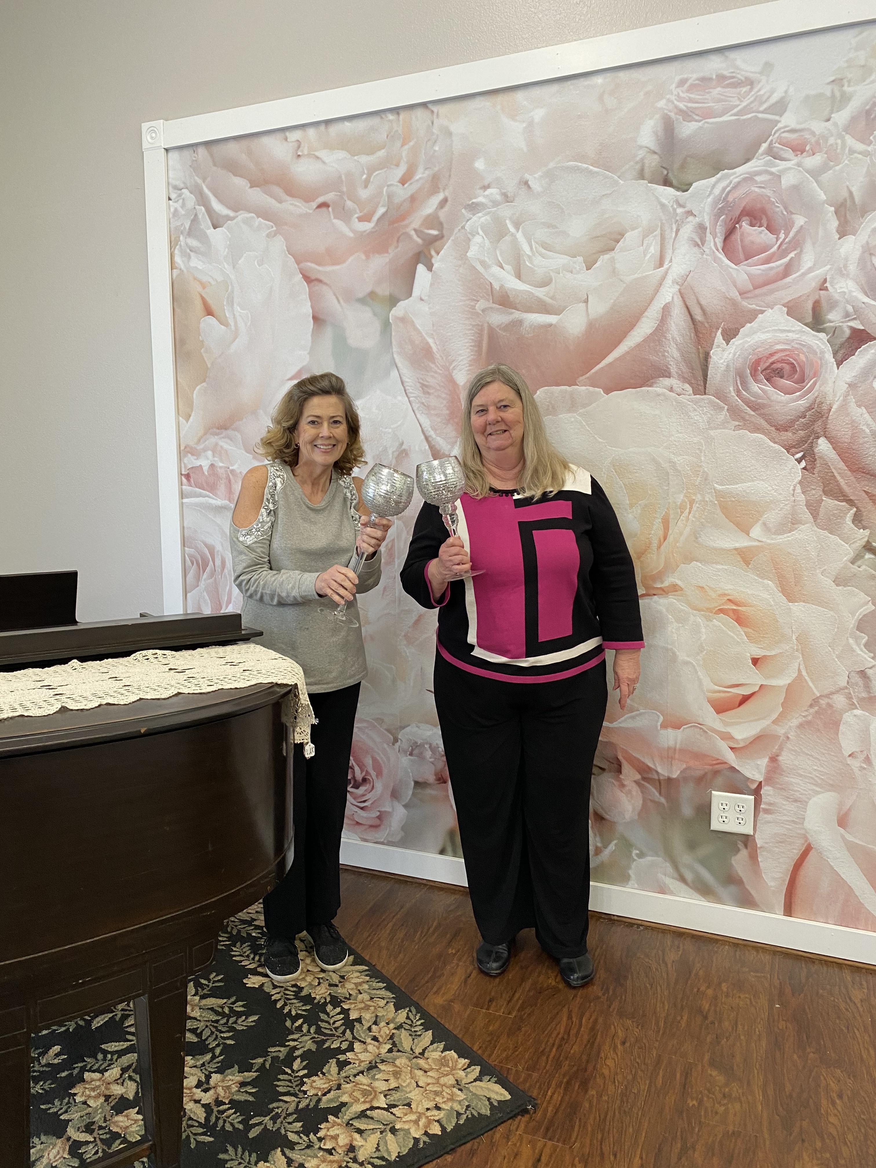Two women holding silver cups in front of a floral wall